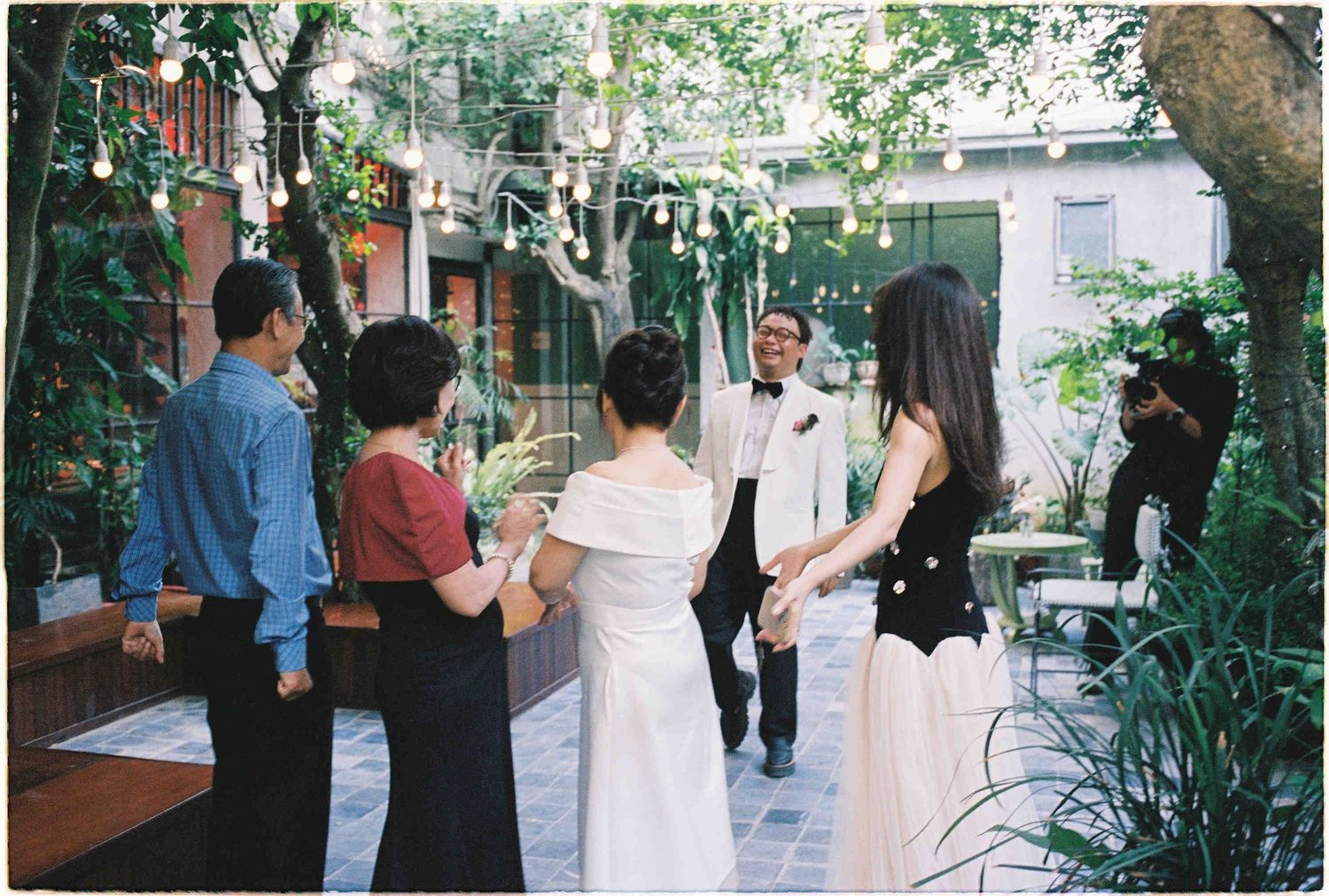 Outdoor wedding ceremony with guests gathering in a lush garden courtyard with string lights and greenery.
