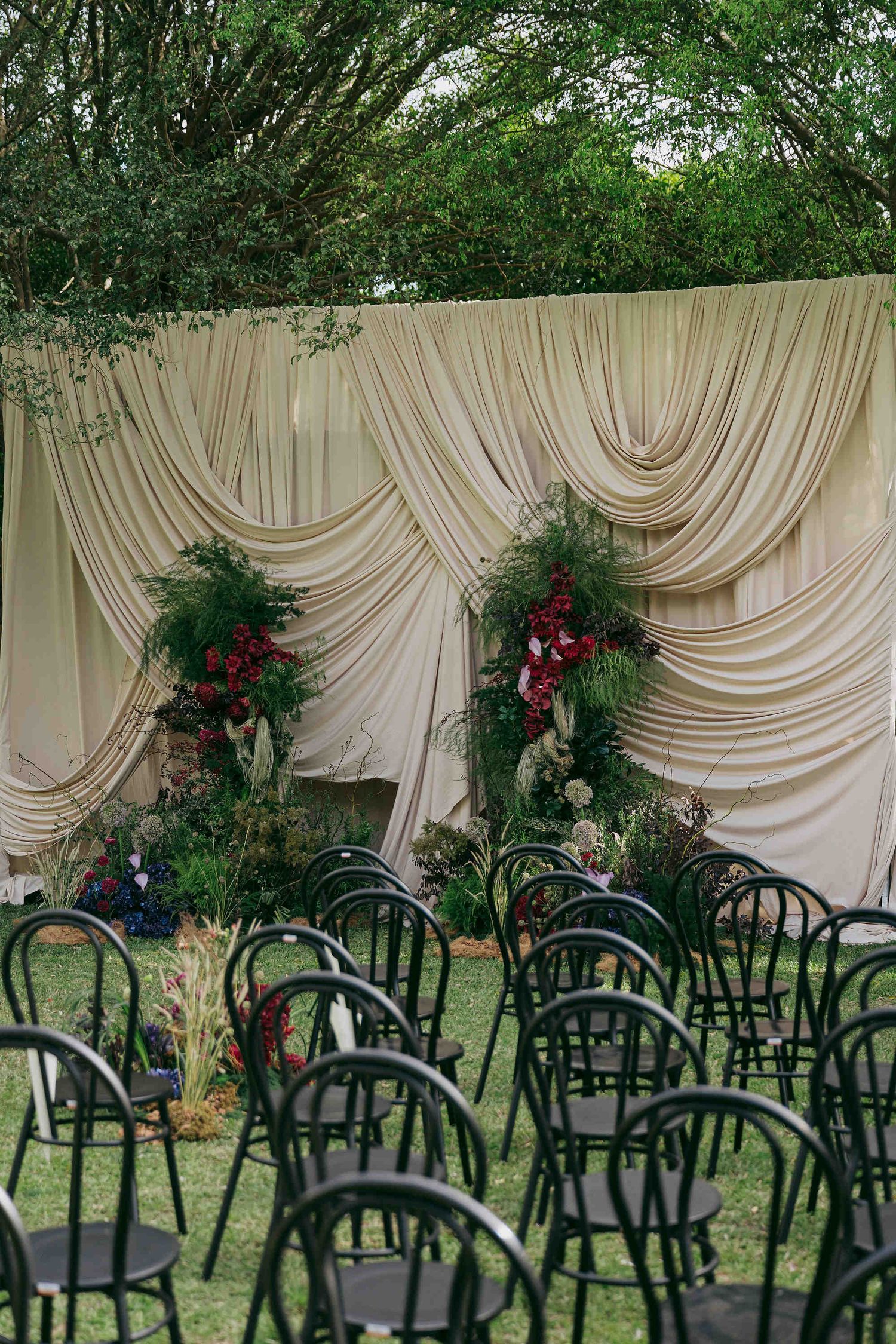 Outdoor wedding ceremony setup with black chairs, draped cream fabric backdrop, and lush red and green floral arrangements on green lawn.