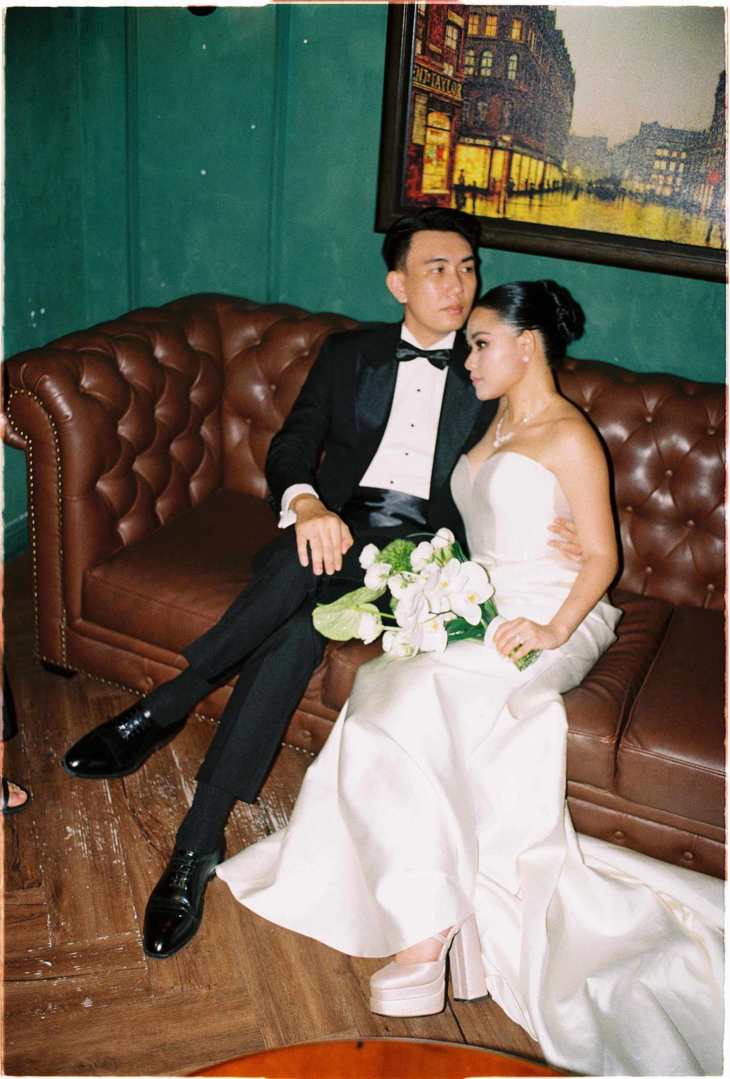 Vietnam pre-wedding: newlywed couple relaxes on brown leather couch during wedding reception, by AnhVuWedding.