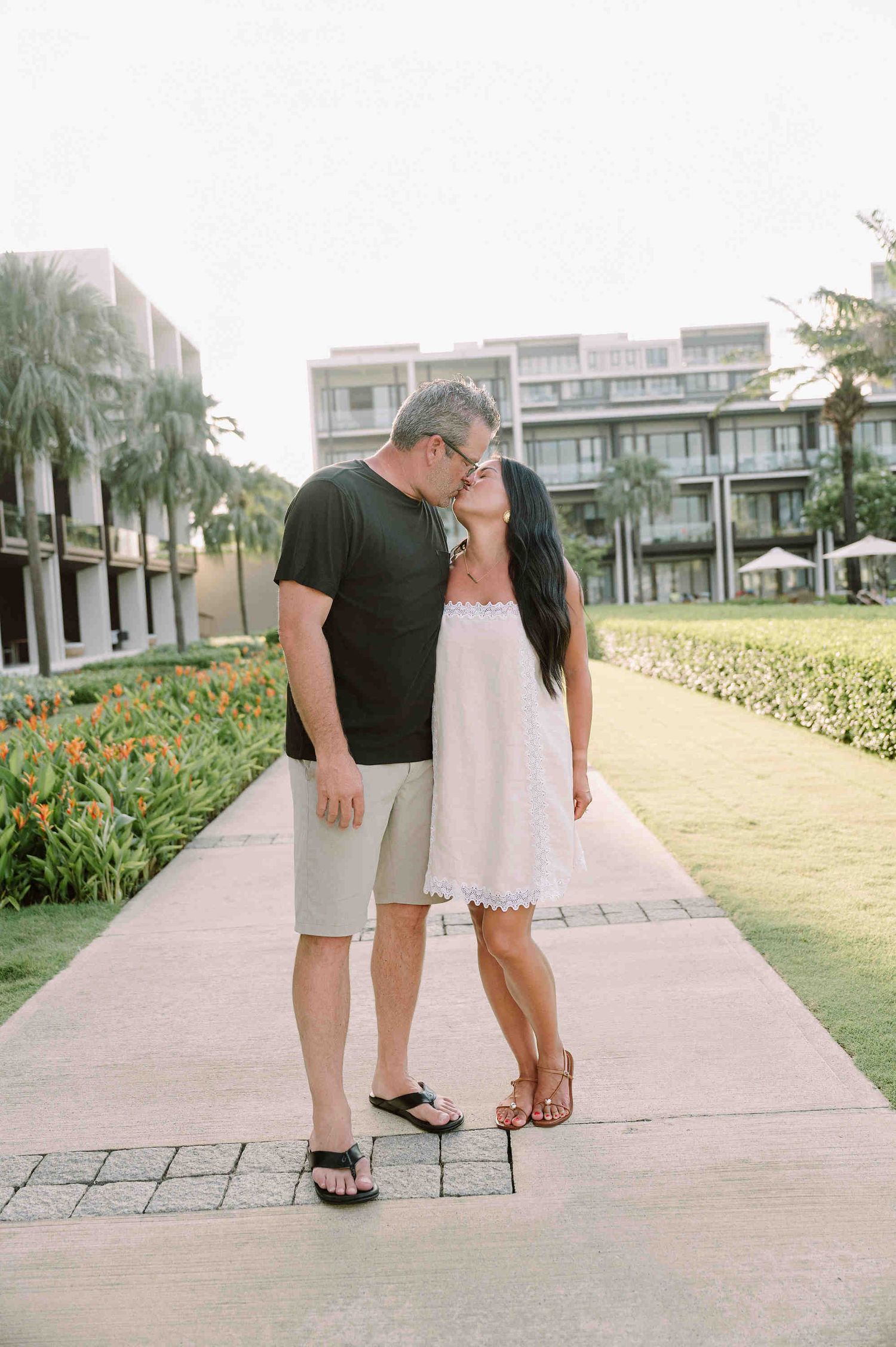 Da Nang family: couple shares a romantic moment on a palm-lined pathway at a tropical resort with landscaped gardens, by AnhVuWedding.
