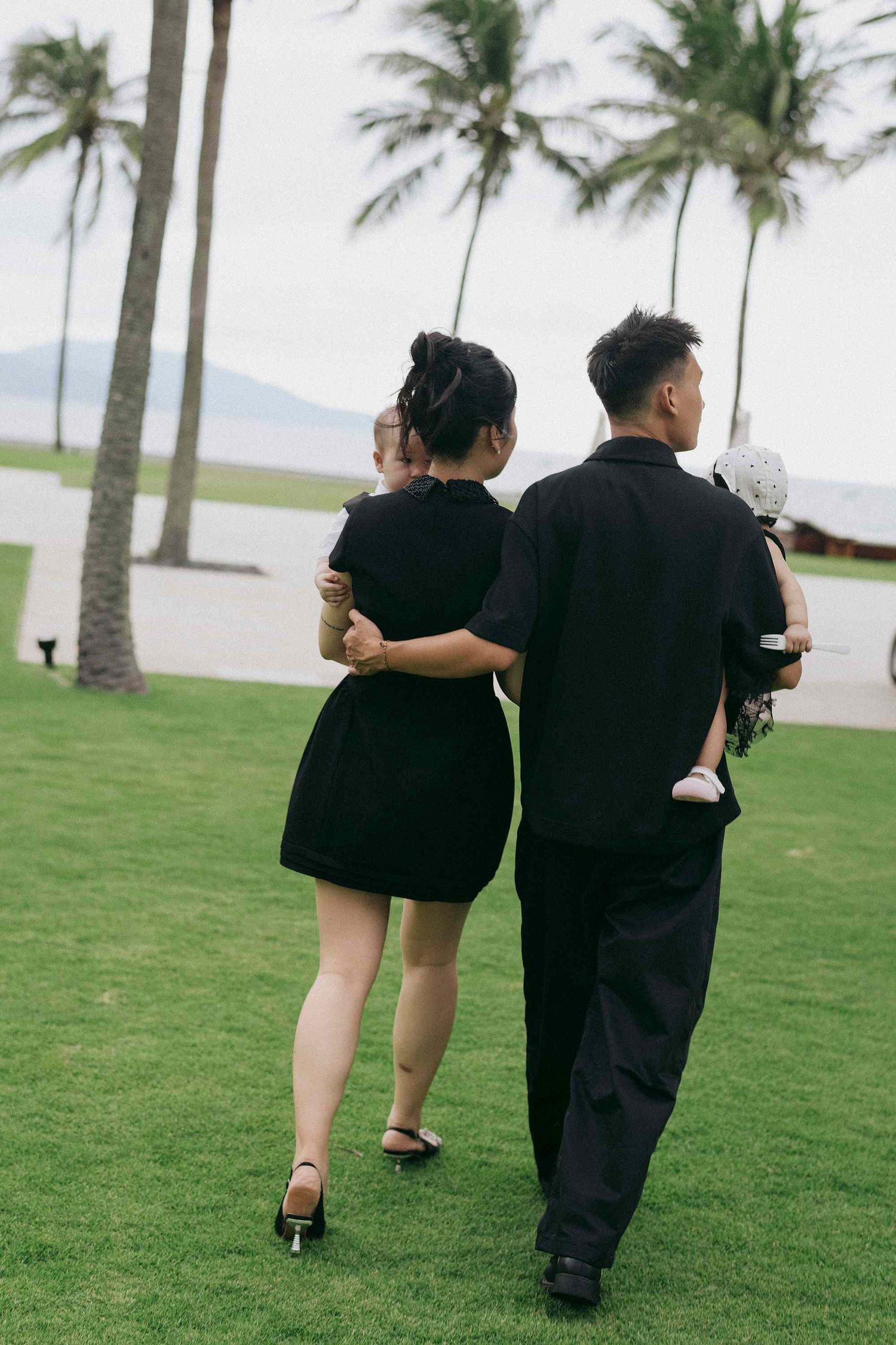 Da Nang family: palm trees line a grassy area where people in black attire stroll together, by AnhVuWedding.