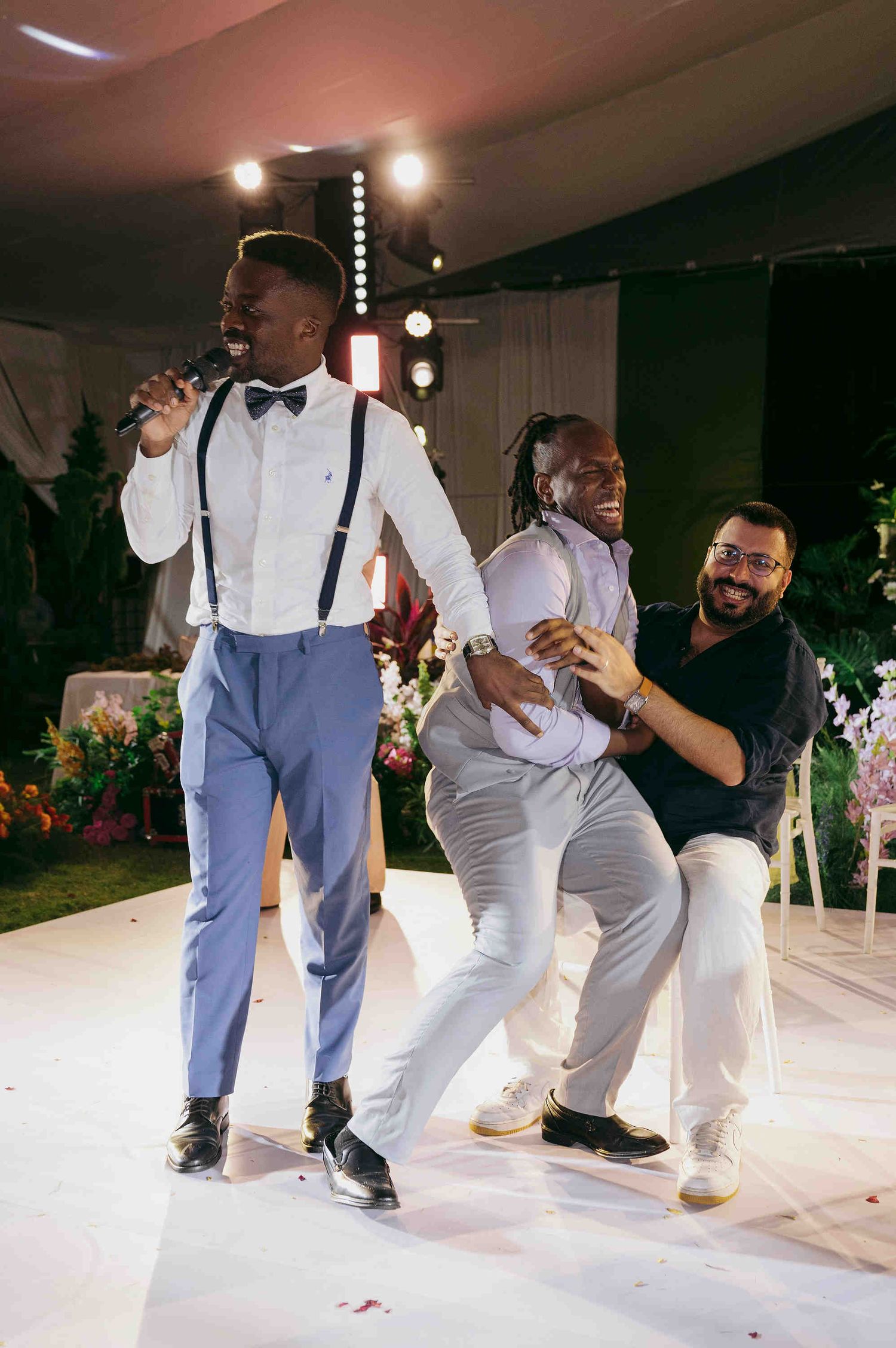 Three men dancing and celebrating on a white dance floor at an elegant indoor event or wedding reception.
