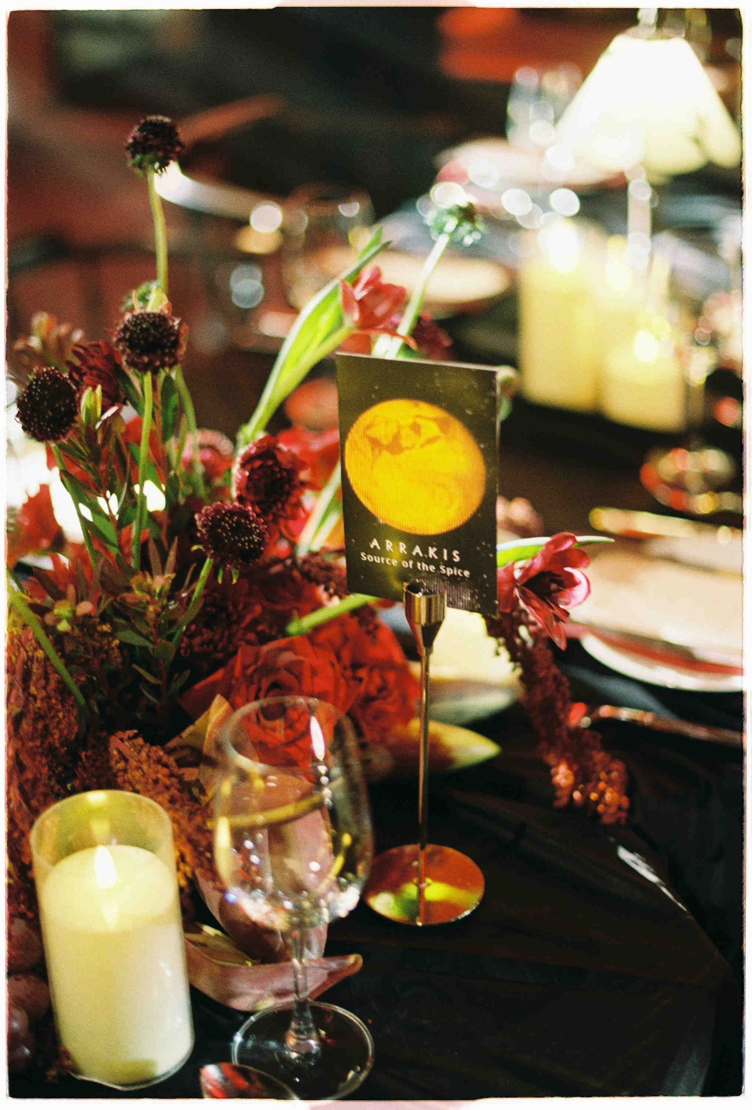 Elegant Dune-themed table setting with Arrakis card, dark florals, candles, and wine glasses on black tablecloth.