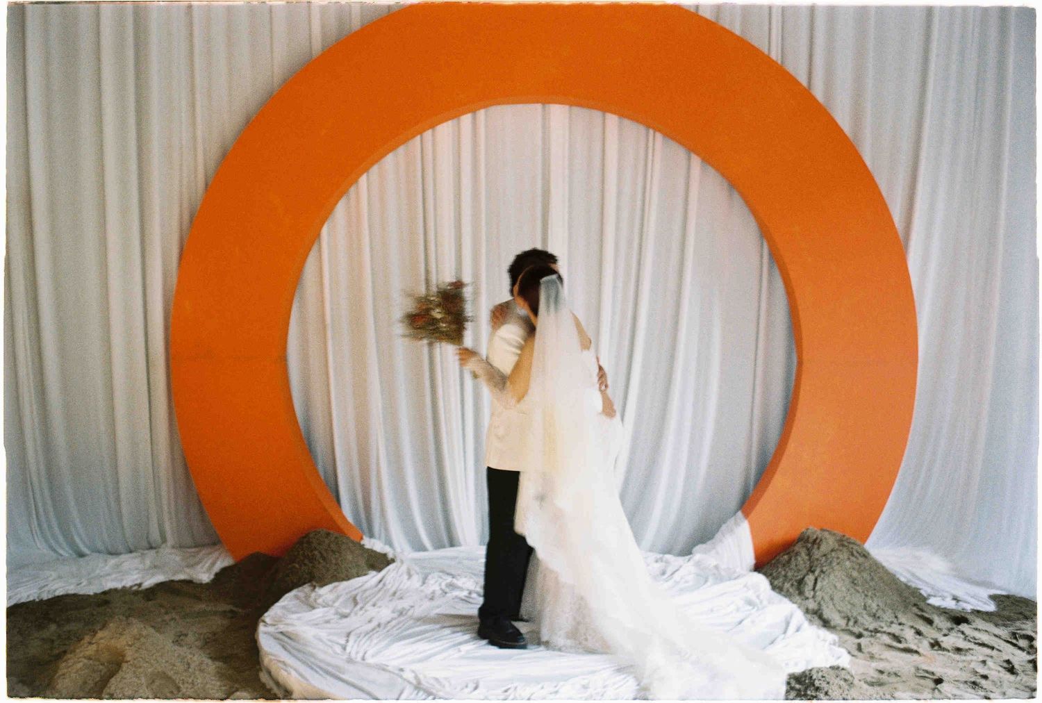 Bride and groom embrace before a large orange circular arch backdrop with white draping at a wedding ceremony.