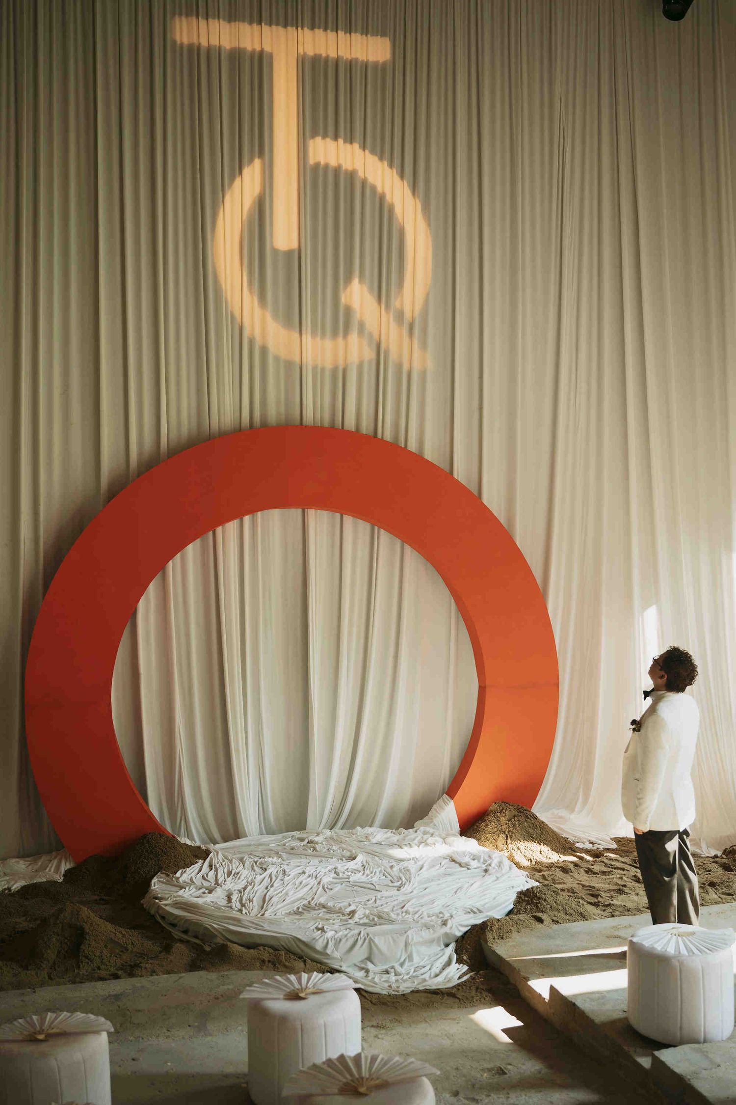 A person stands beside a large orange circular arch installation against white drapes with a projected TQ logo above.