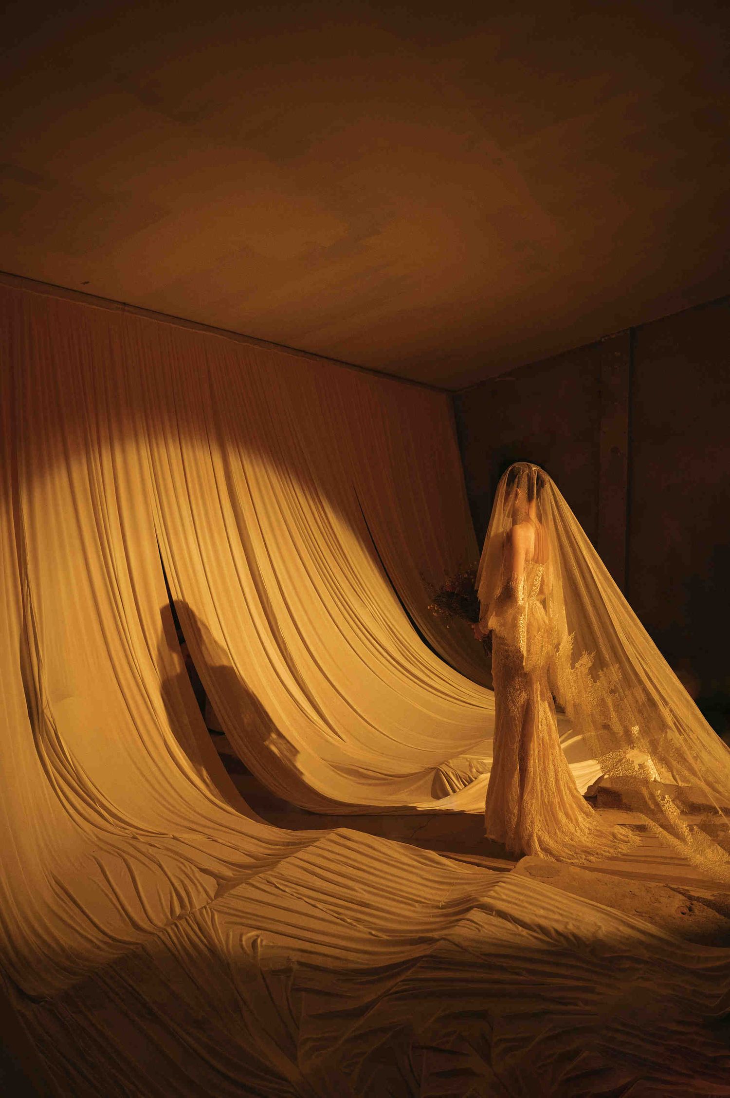 A figure draped in flowing white fabric stands in a warmly lit room surrounded by billowing curtains.