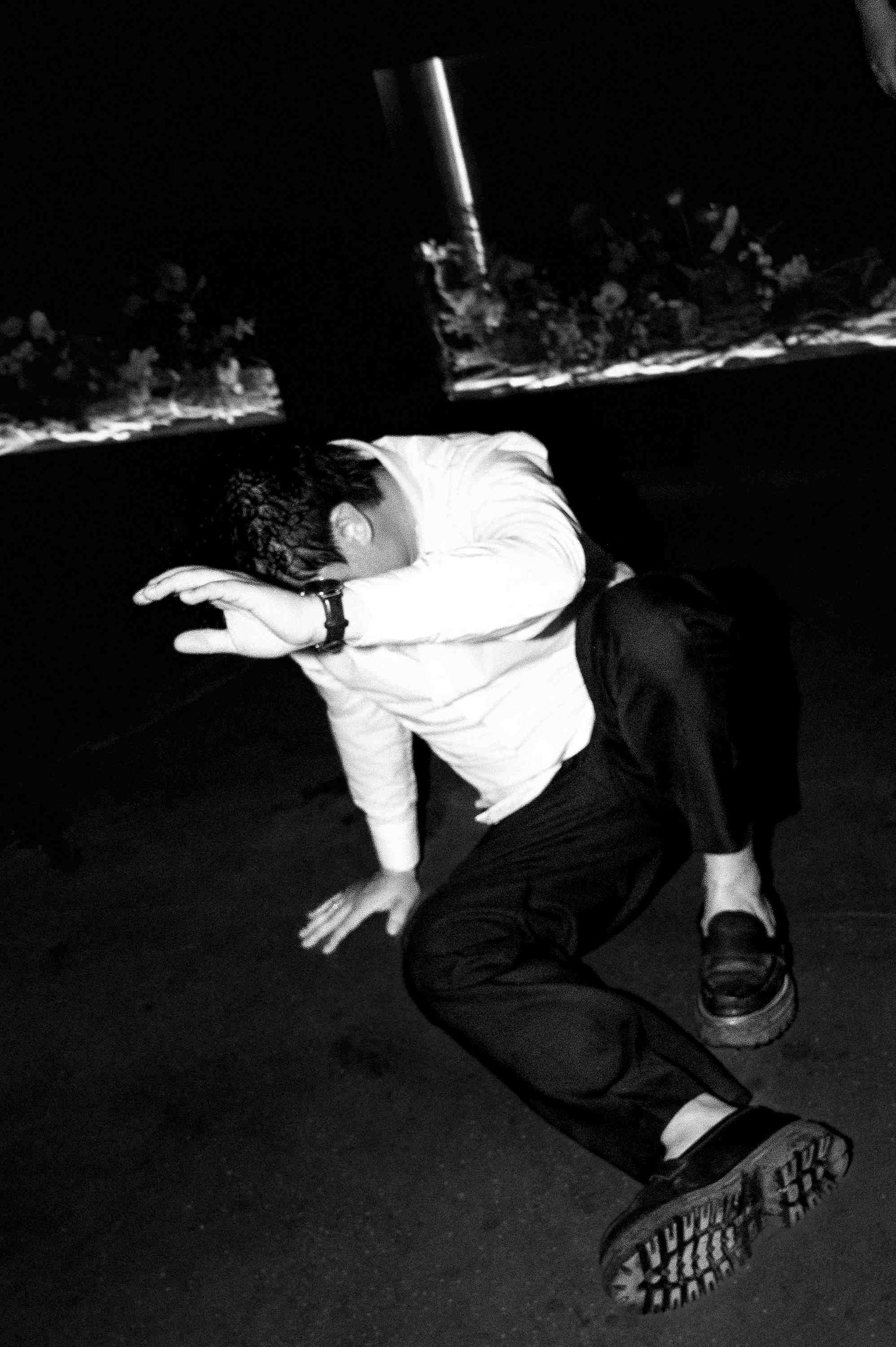 Black and white photo of a man in white shirt and black pants crouching low in a dynamic dance or street performance pose.
