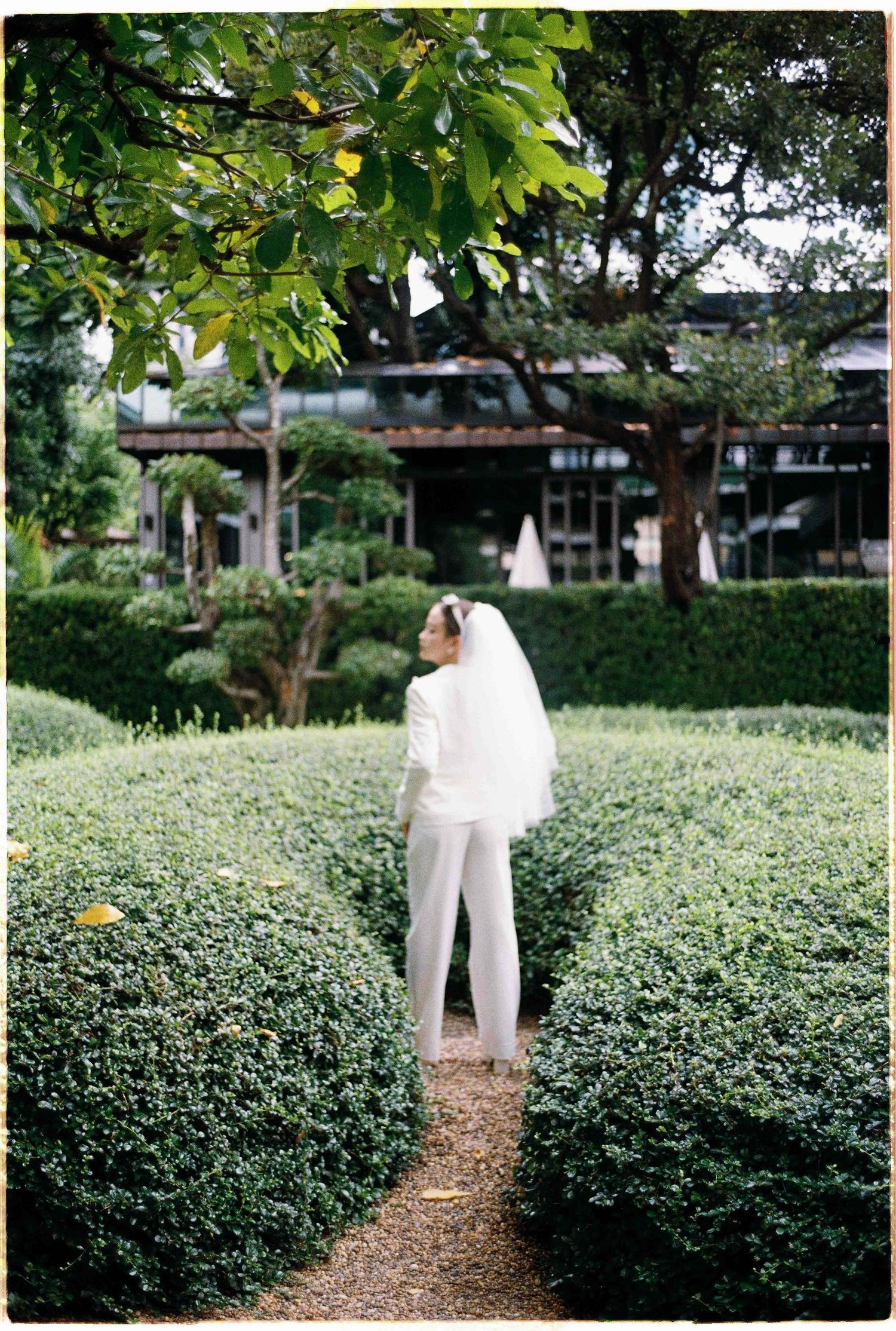 Saigon pre-wedding: figure in white dress walking through a garden path lined with green hedges, by AnhVuWedding.