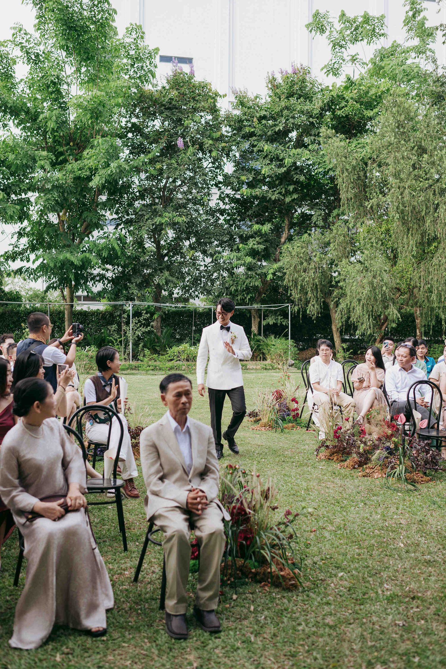 Outdoor garden wedding ceremony with guests seated on chairs, groom in white jacket walking down the aisle.