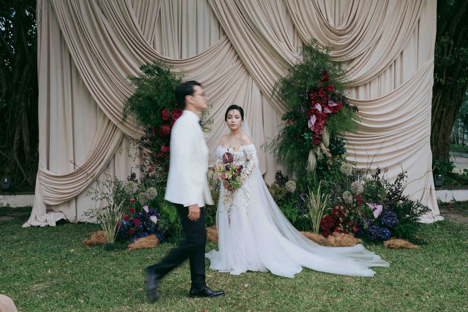 Bride in white gown and groom in black suit stand before draped fabric backdrop with lush floral arrangements at outdoor wedding.