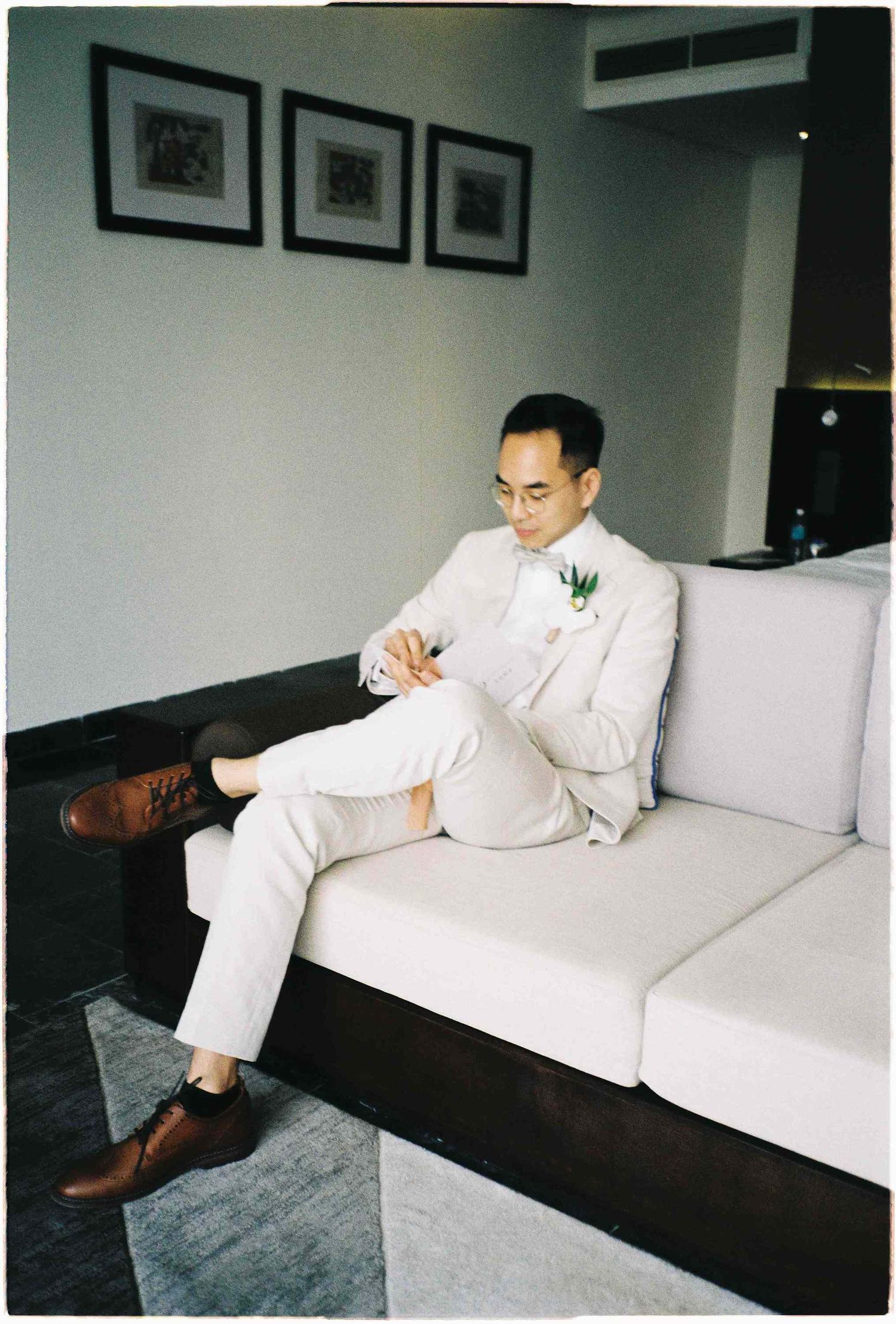 Groom in white suit with boutonniere sits on modern sofa, adjusting cufflinks before wedding ceremony.