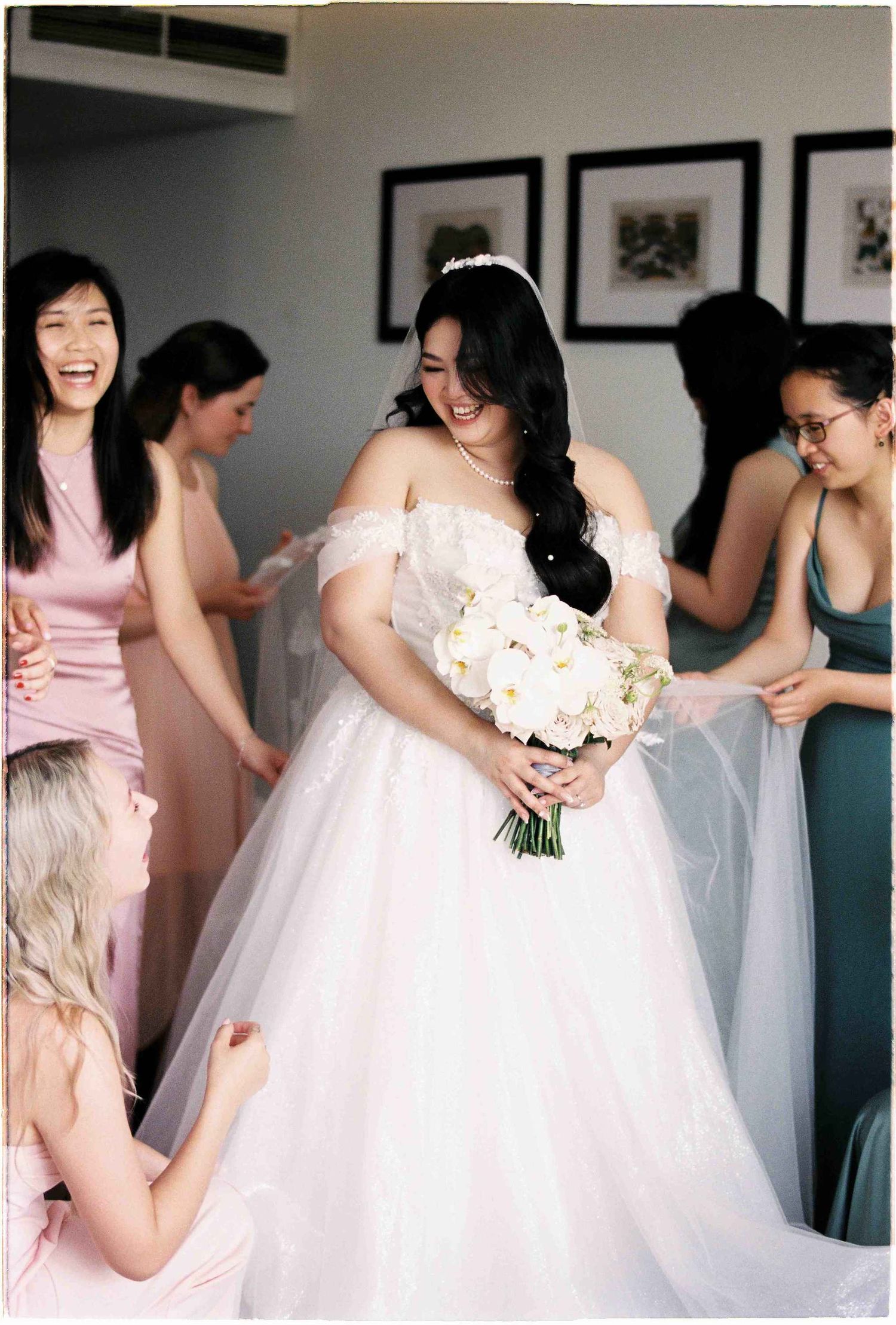 Bride in white off-shoulder ball gown holding white bouquet surrounded by bridesmaids in pink and green dresses.