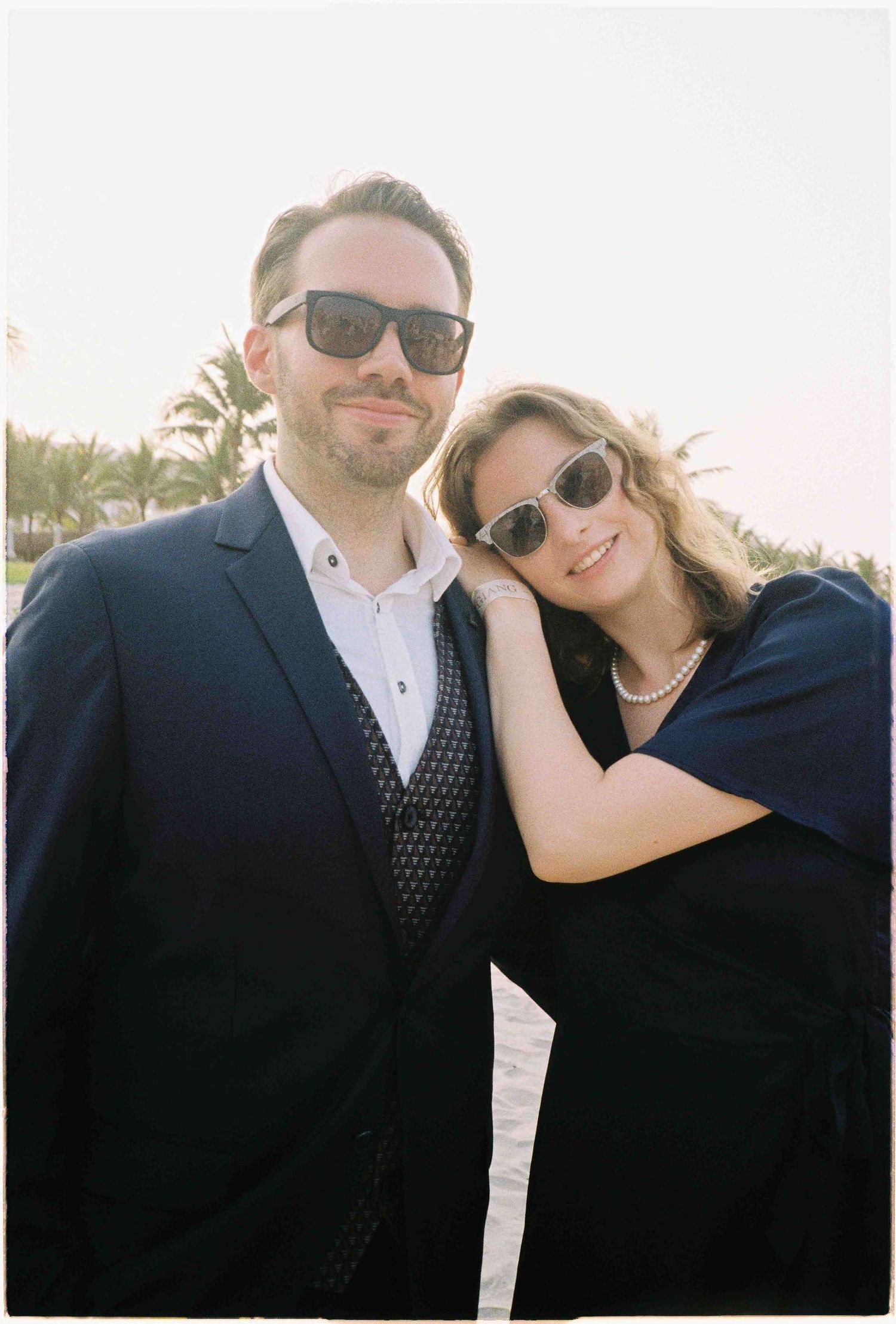 Couple in sunglasses and navy attire posing together on a sunny beach with palm trees in the background.