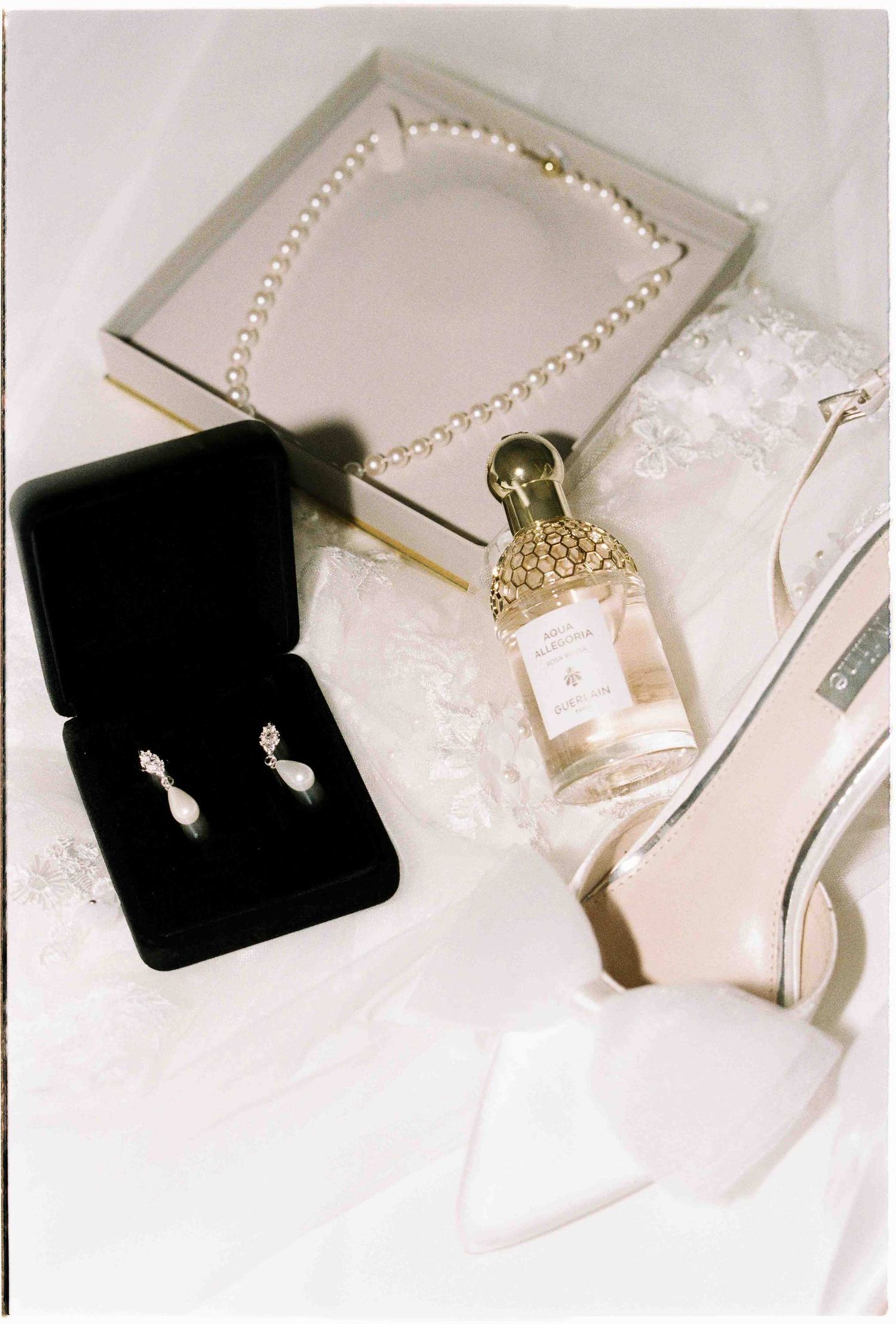 Elegant bridal accessories flat lay with pearl necklace, drop earrings, Guerlain perfume, and ivory heels.