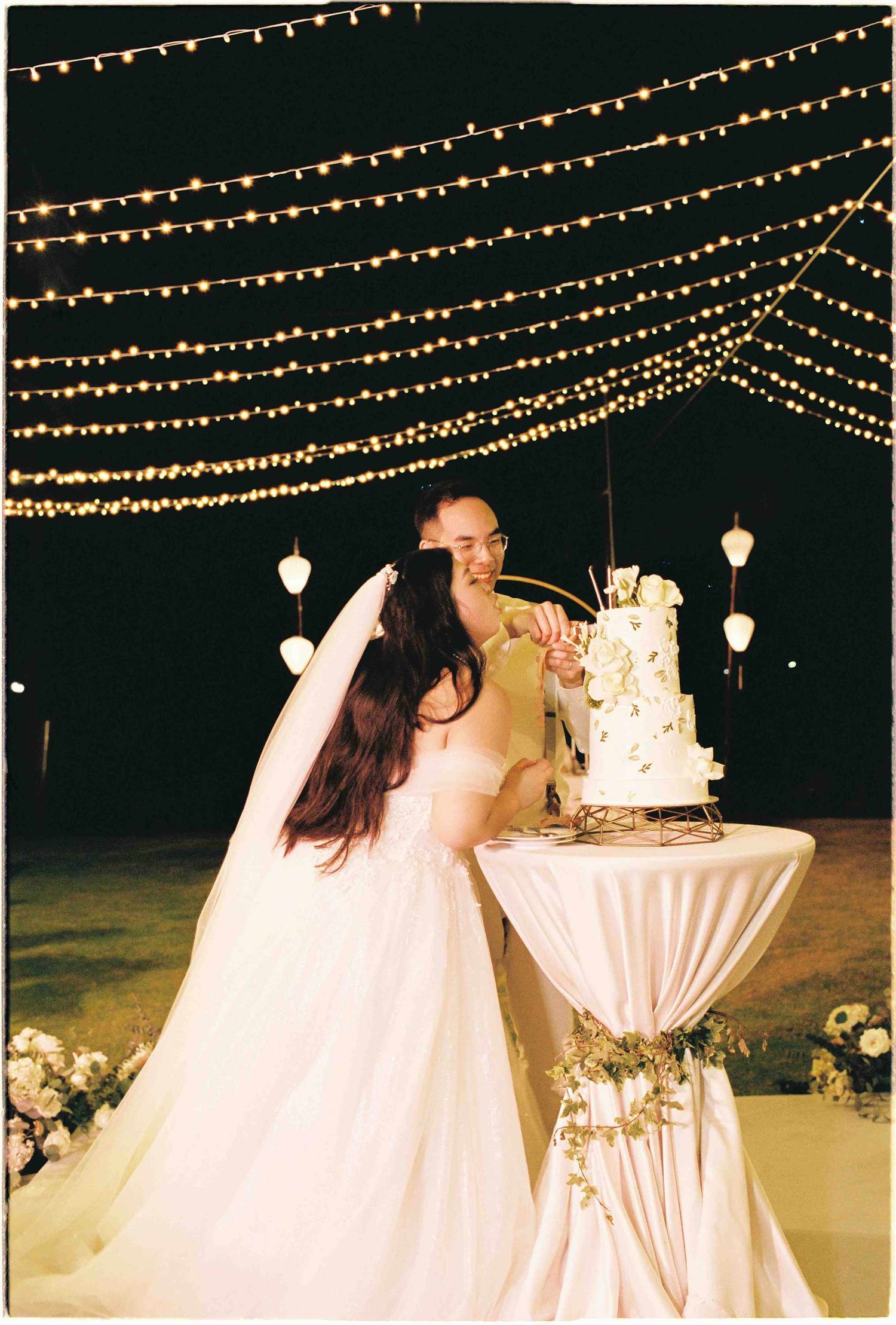 Bride and groom cutting wedding cake outdoors at night under romantic string lights at elegant reception.