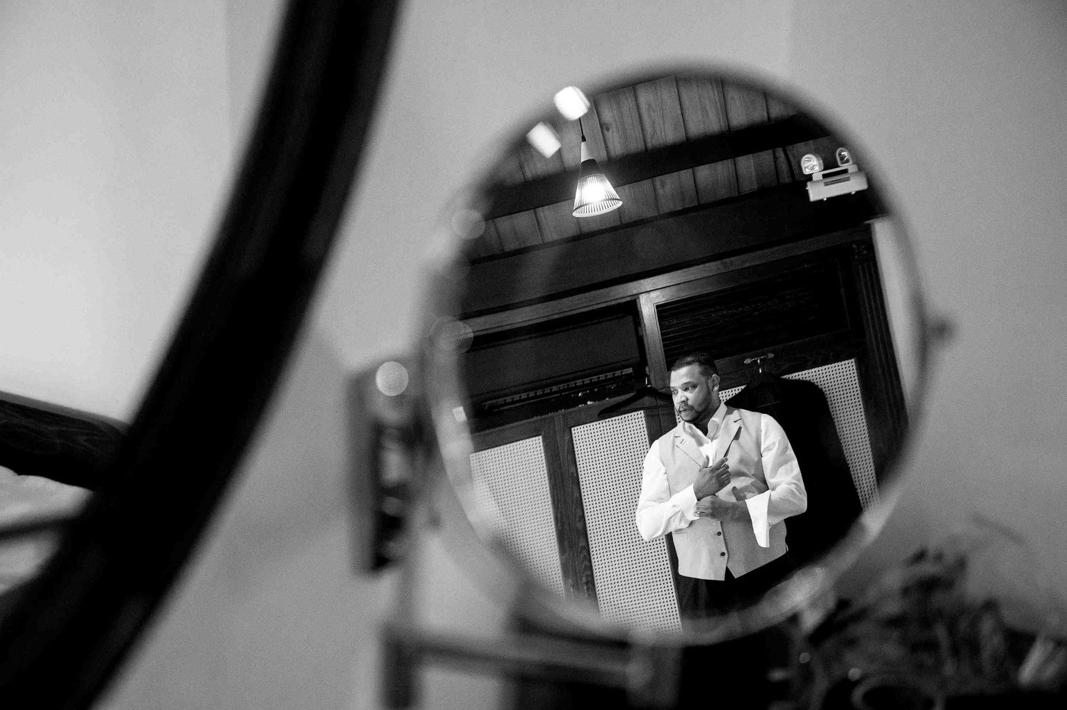 Black and white mirror reflection of a man in formal attire buttoning his shirt before a wedding ceremony.