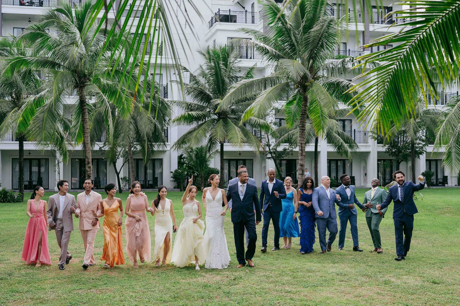 Wedding party walks together on tropical lawn with palm trees and white resort building backdrop.