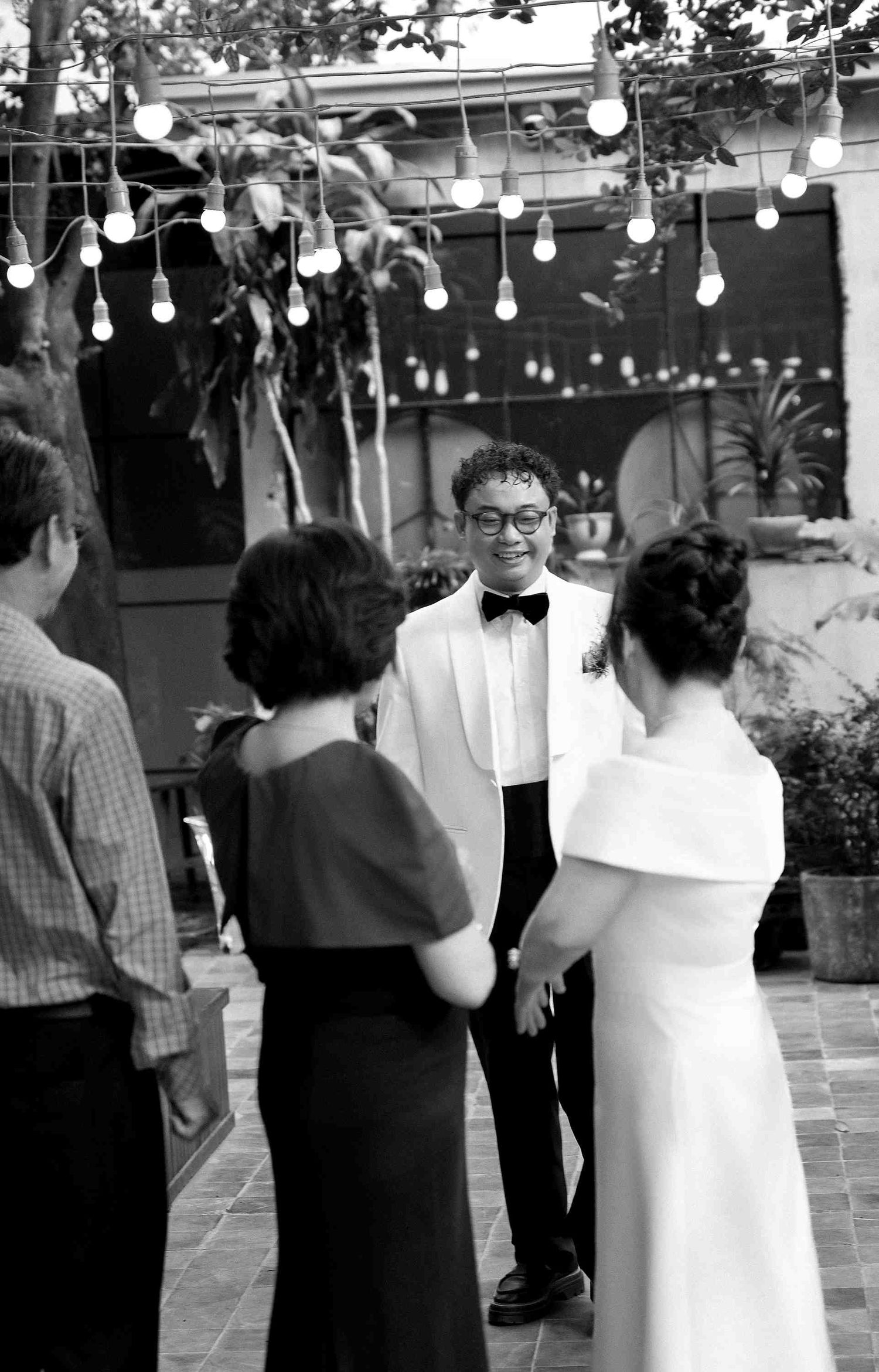 Black and white photo of a groom in white tuxedo holding hands with bride at an outdoor wedding ceremony.