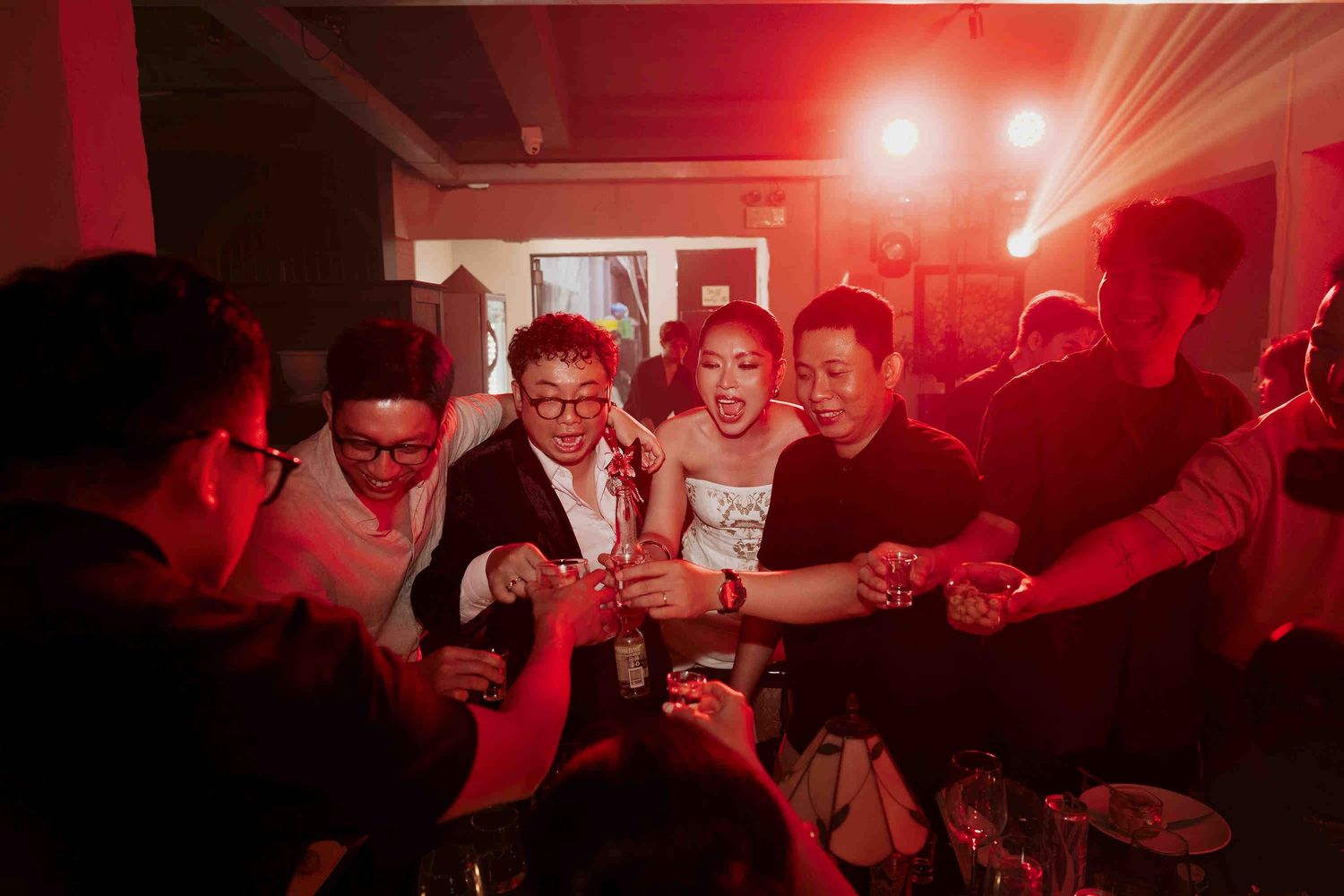 Group of friends celebrating at a nightclub party under red lights, raising drinks in a toast together.