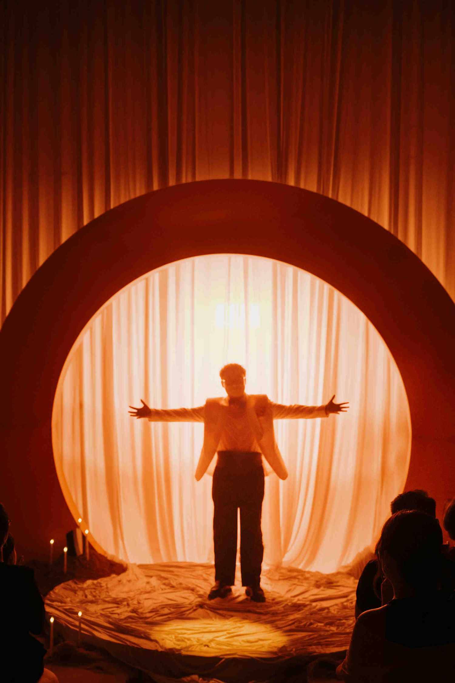 Silhouette of a person with arms outstretched standing in a circular arch with glowing white curtains on a warm red stage.
