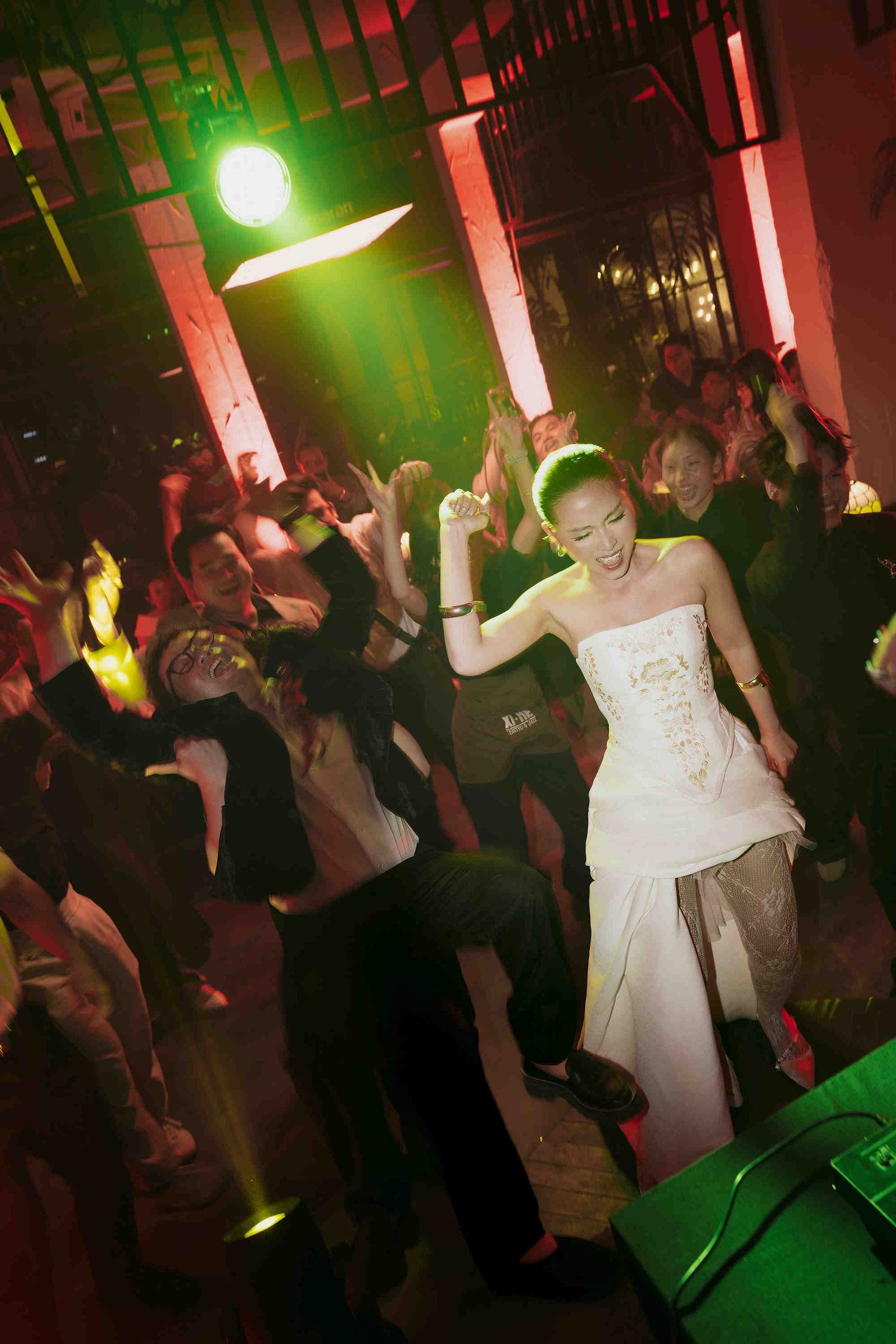 Bride in white strapless gown dancing joyfully at a lively wedding reception with guests under green stage lights.