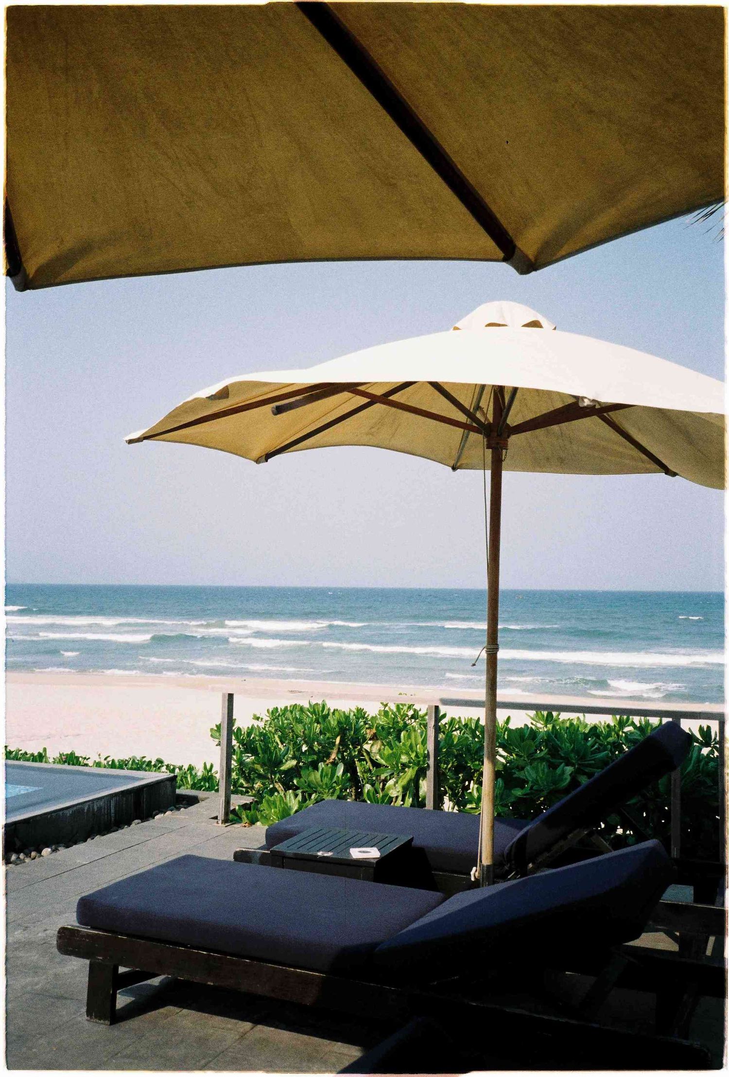 Luxury beach lounge chairs with dark cushions under white patio umbrella overlooking serene ocean waves and sandy shoreline.