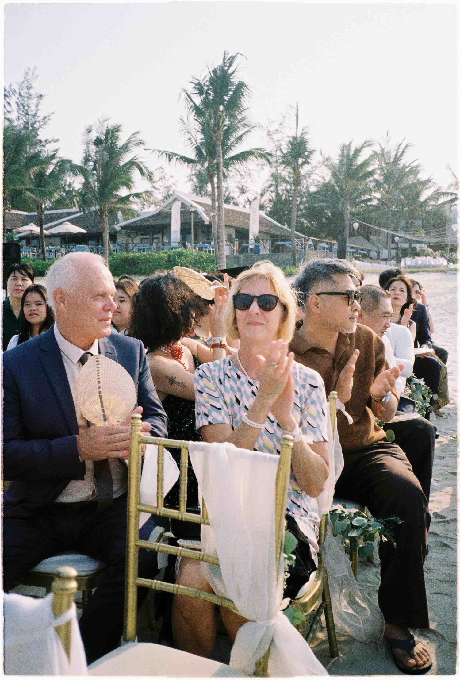 Guests clapping and smiling at an outdoor beach wedding ceremony with palm trees in the background.