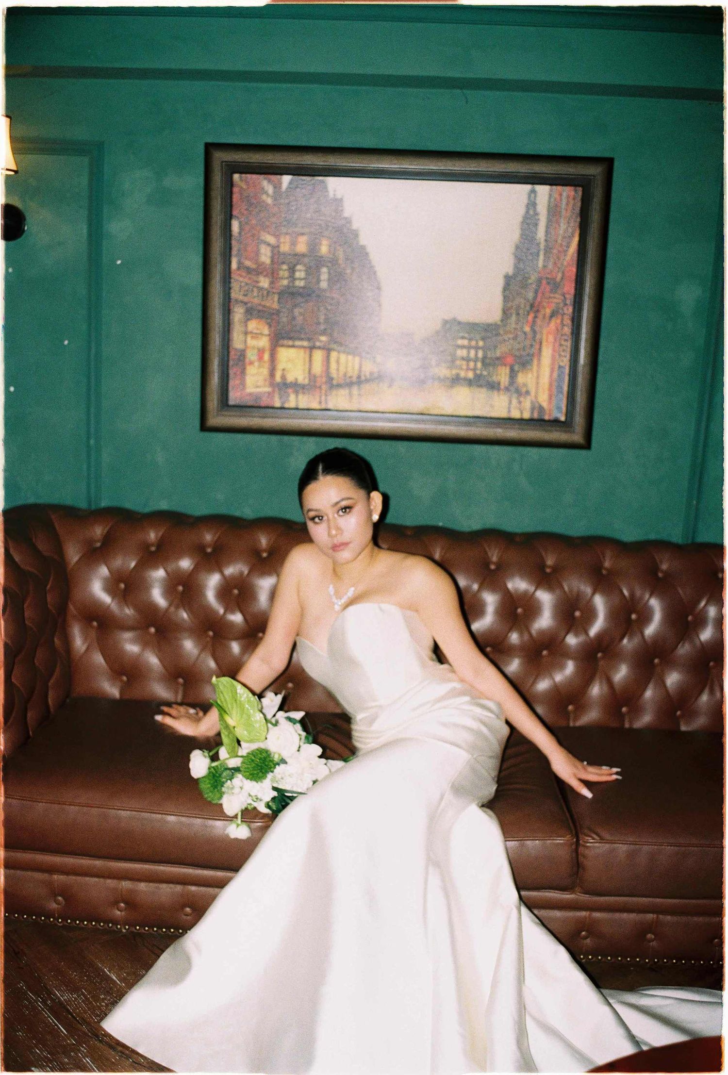 Vietnam pre-wedding: bride in fitted white gown poses on brown leather couch with cityscape artwork in background, by AnhVuWedding.
