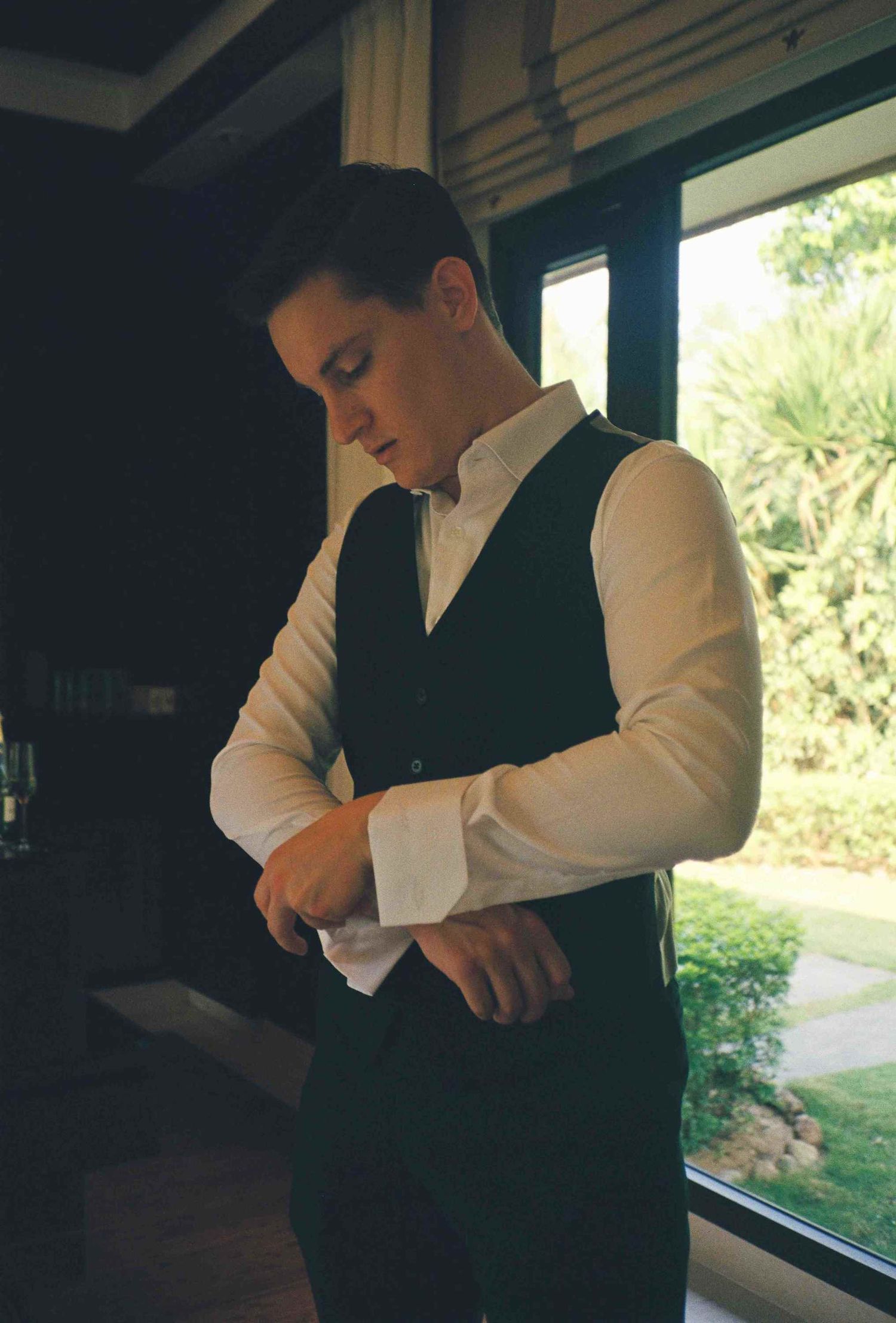 Lang Co wedding: person adjusting formal attire while standing by a window in natural light, by AnhVuWedding.
