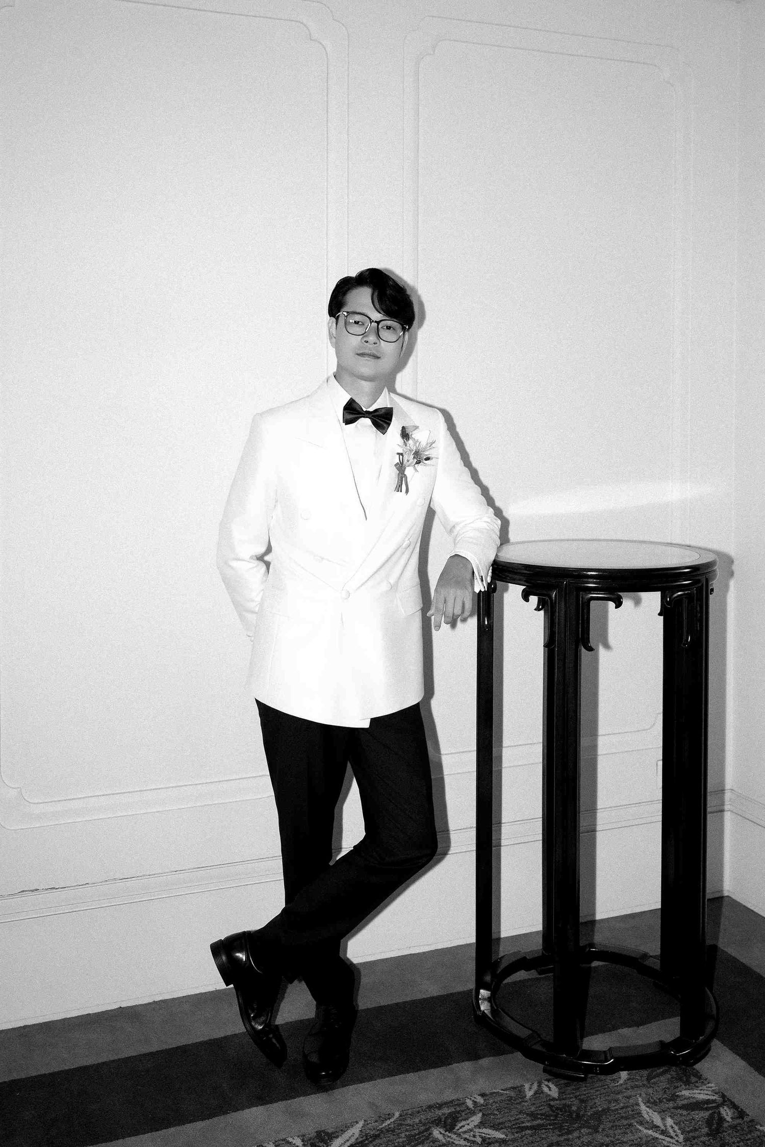 A man in a white tuxedo jacket and black bow tie leans against a pedestal table in a black and white photo.