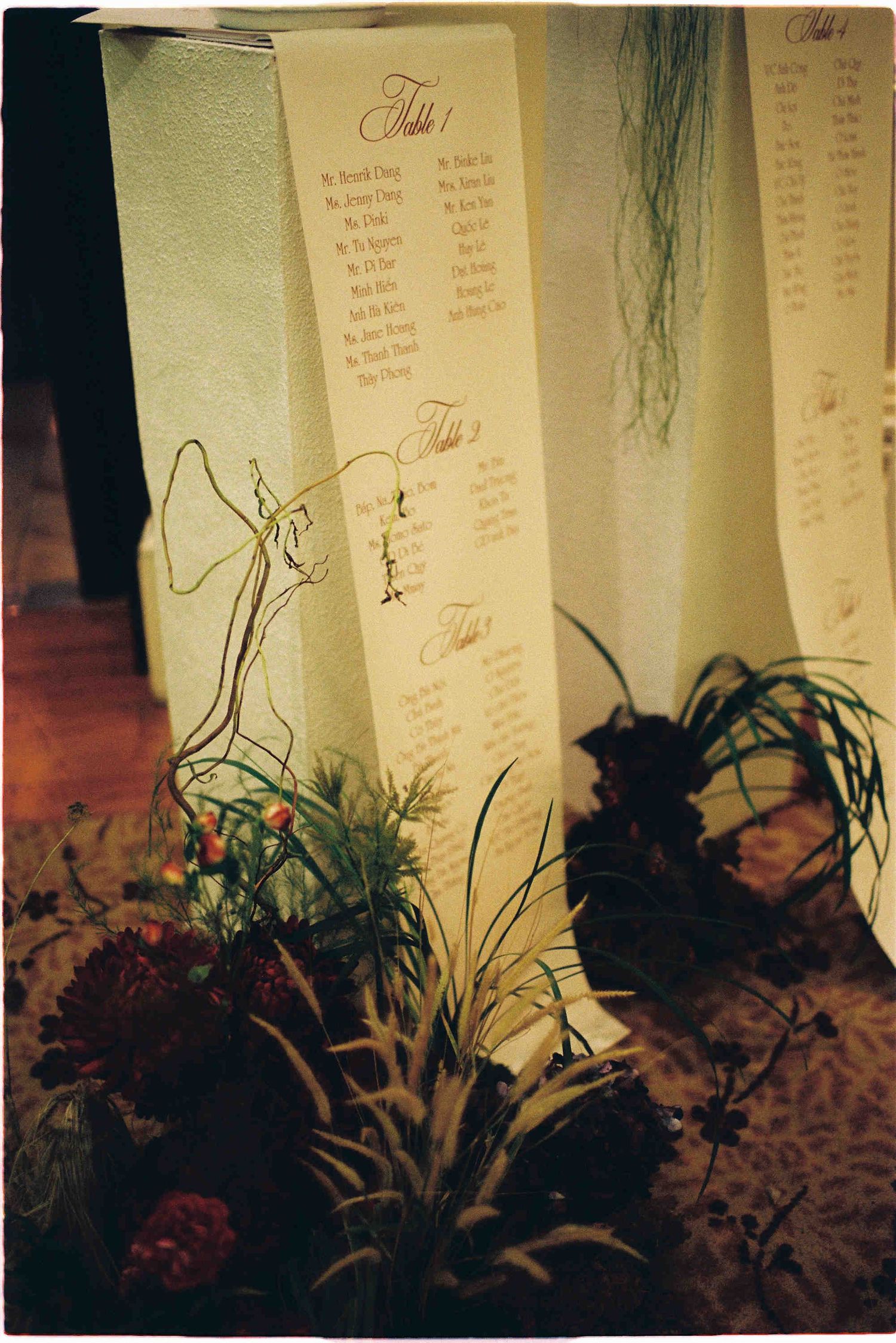 Elegant wedding seating chart displayed on tall cream cards with calligraphy, surrounded by lush green plants and floral arrangements.