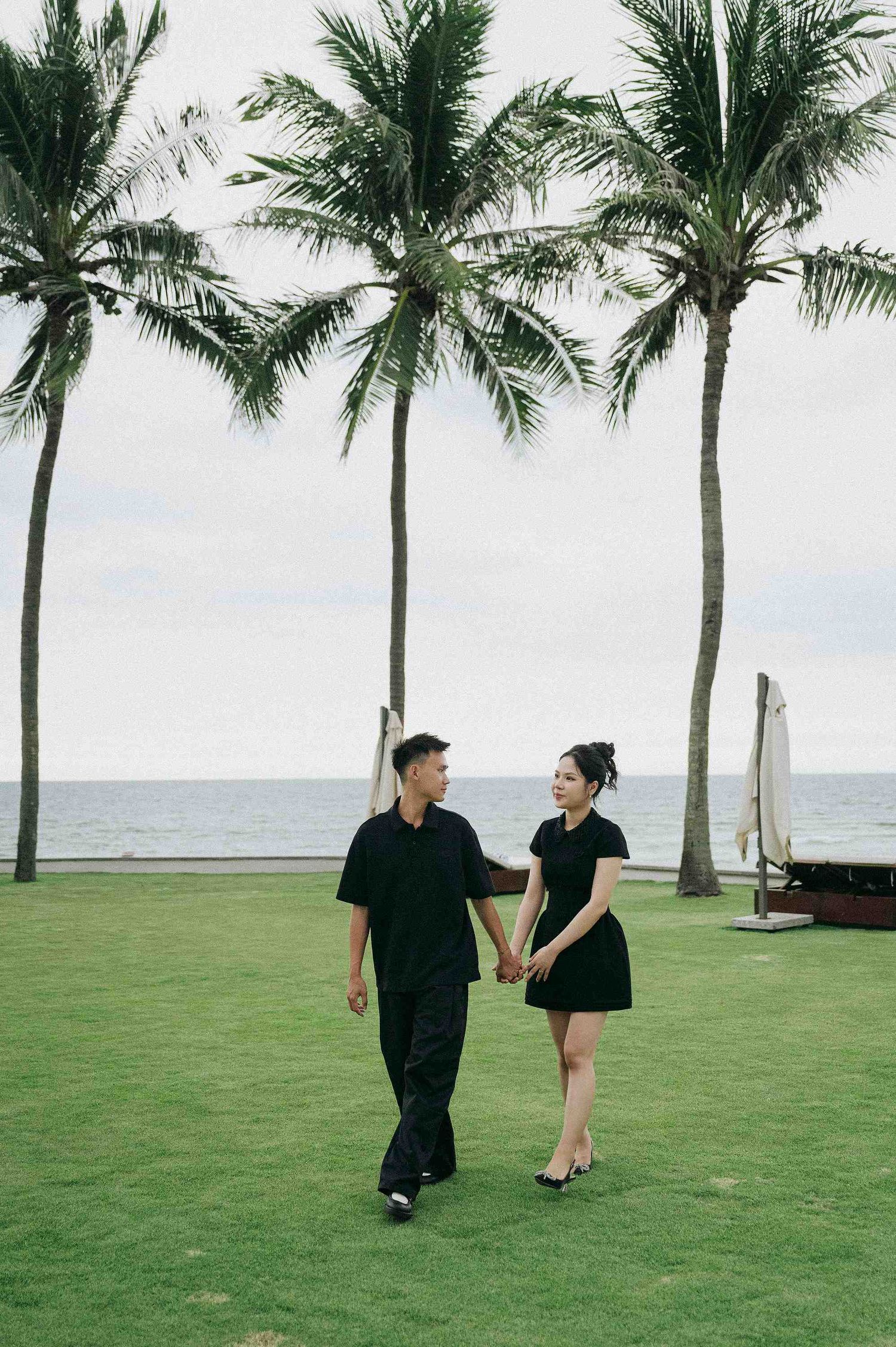 Da Nang family: couple in black outfits walks along a palm-lined beachfront lawn on a sunny day, by AnhVuWedding.