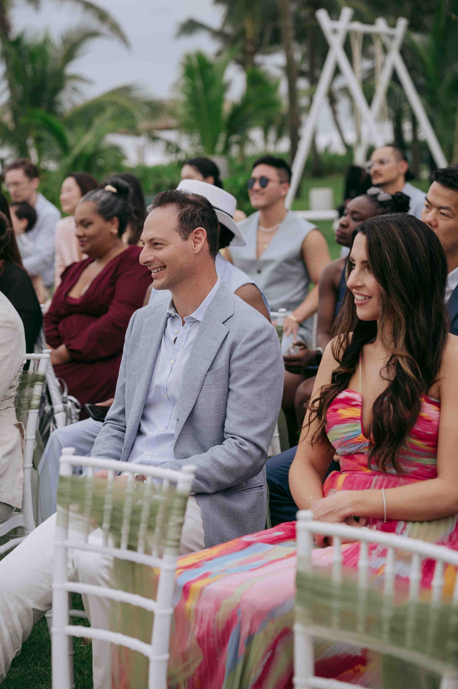 Guests smiling and laughing at an outdoor wedding ceremony, dressed in colorful attire with tropical palm trees in the background.