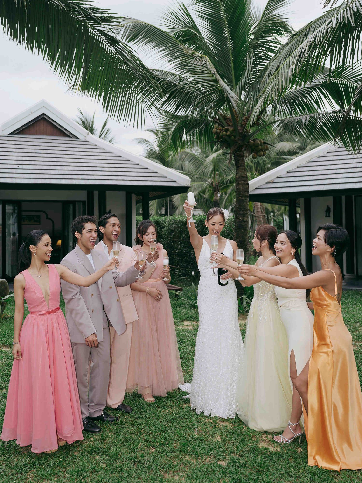 Wedding party toasting with champagne outdoors, bride in white gown with bridesmaids in pink and gold dresses near tropical palm trees.