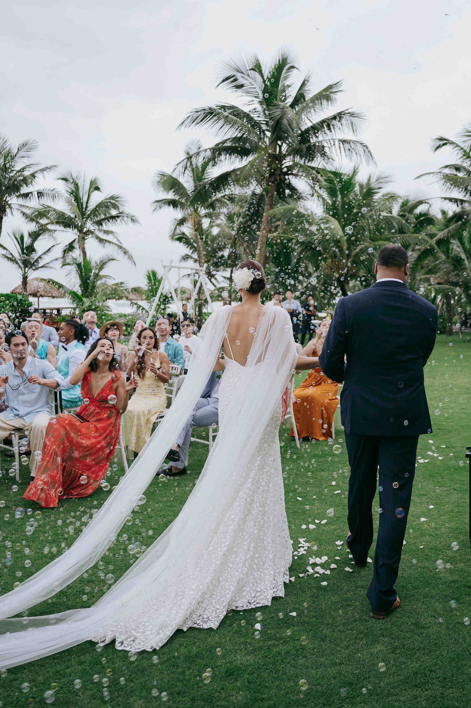 Bride and groom walk down outdoor aisle with long veil, guests cheering, palm trees and bubbles at tropical wedding.