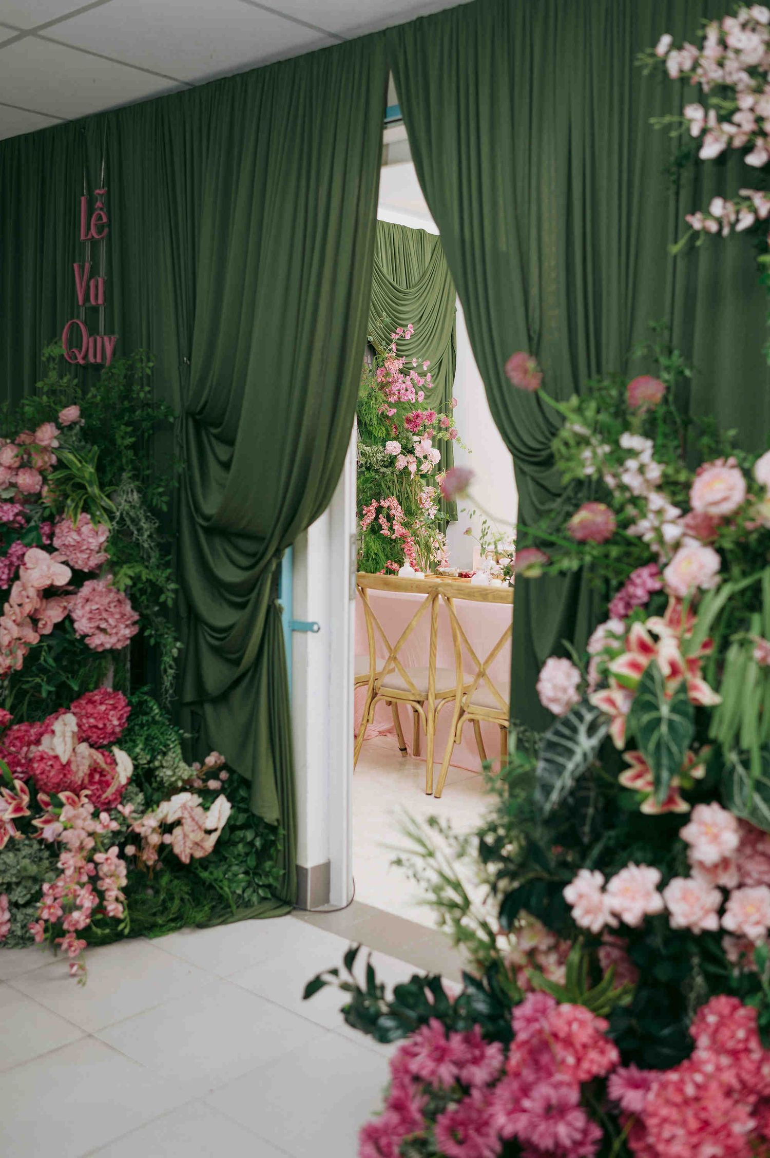 Elegant wedding backdrop with green drapes, pink floral wall, and gold chairs visible through draped doorway.