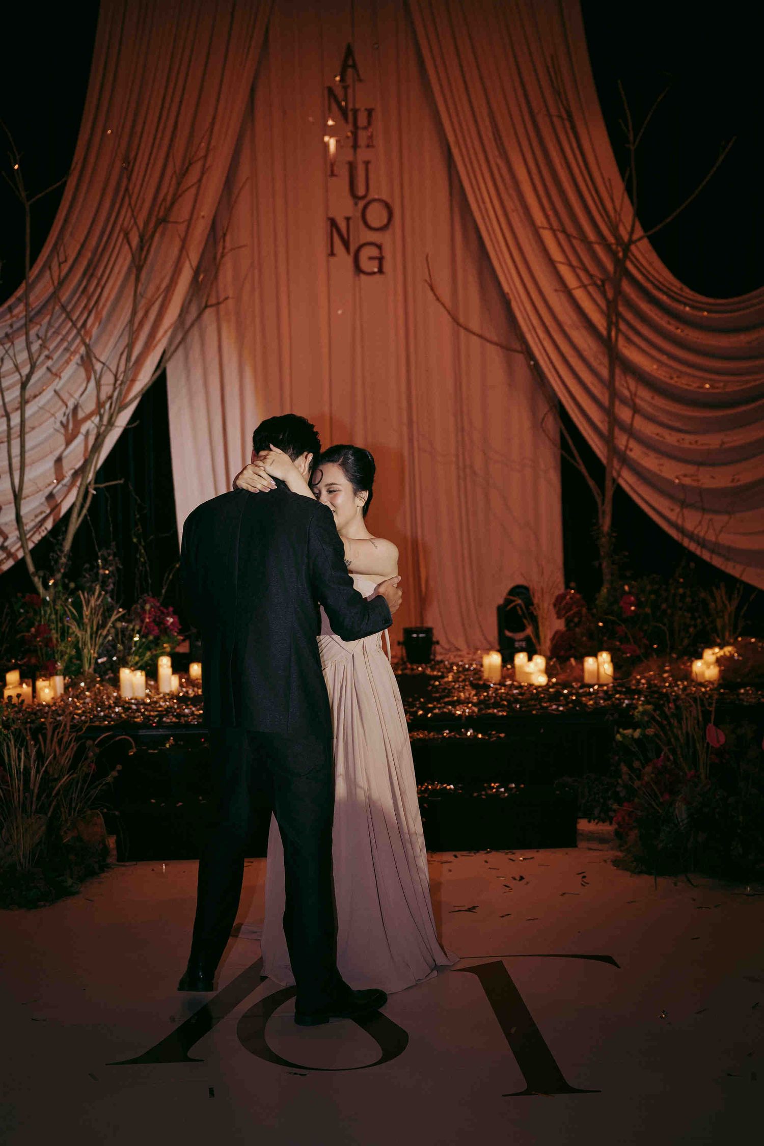 Couple shares first dance at romantic candlelit wedding reception with draped coral backdrop and floral decor.