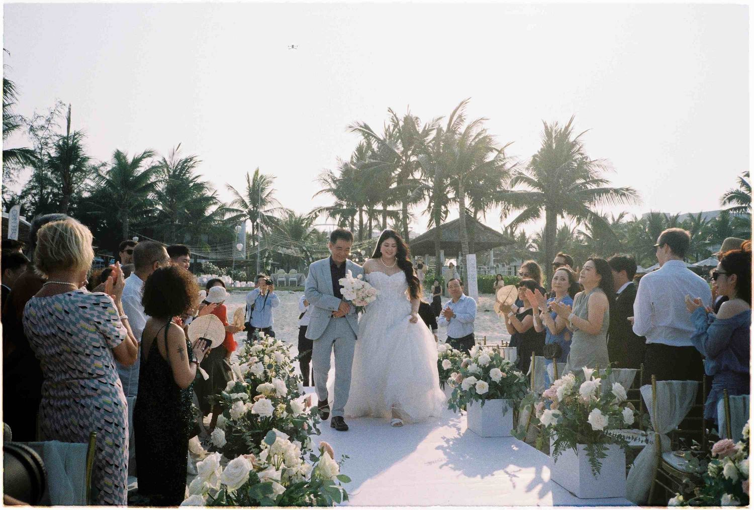 Newlyweds walk down aisle at tropical beach wedding surrounded by guests and white floral arrangements.