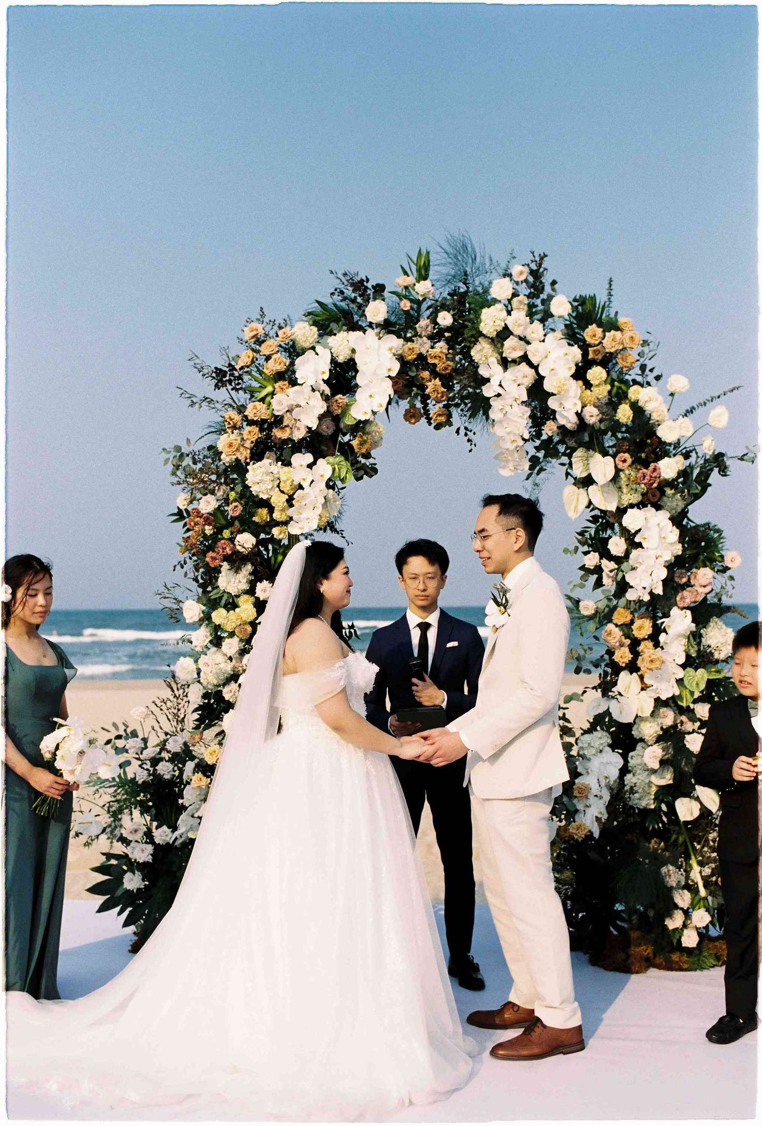 Couple exchanges vows at beach wedding under floral arch with white and yellow blooms, officiant and bridesmaid in background.
