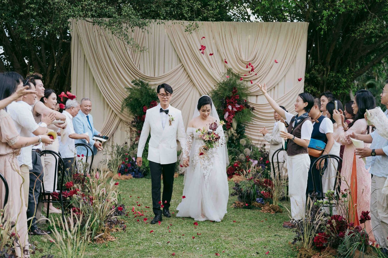 Bride and groom walk down outdoor aisle lined with guests tossing petals, elegant cream floral backdrop.
