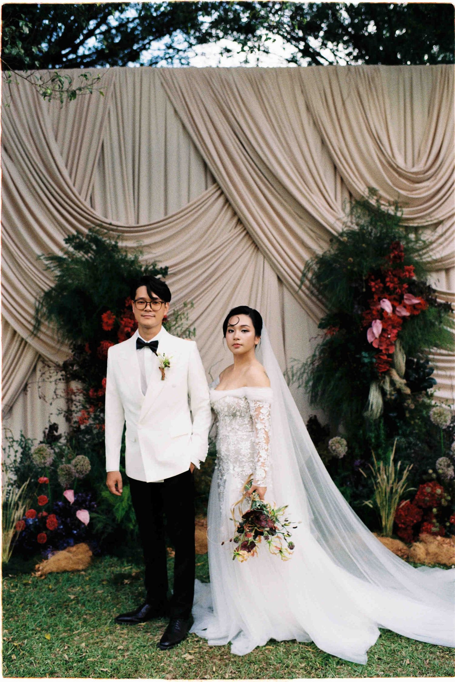 Couple posing in wedding attire before floral backdrop, groom in white tuxedo, bride in lace gown with veil holding bouquet.
