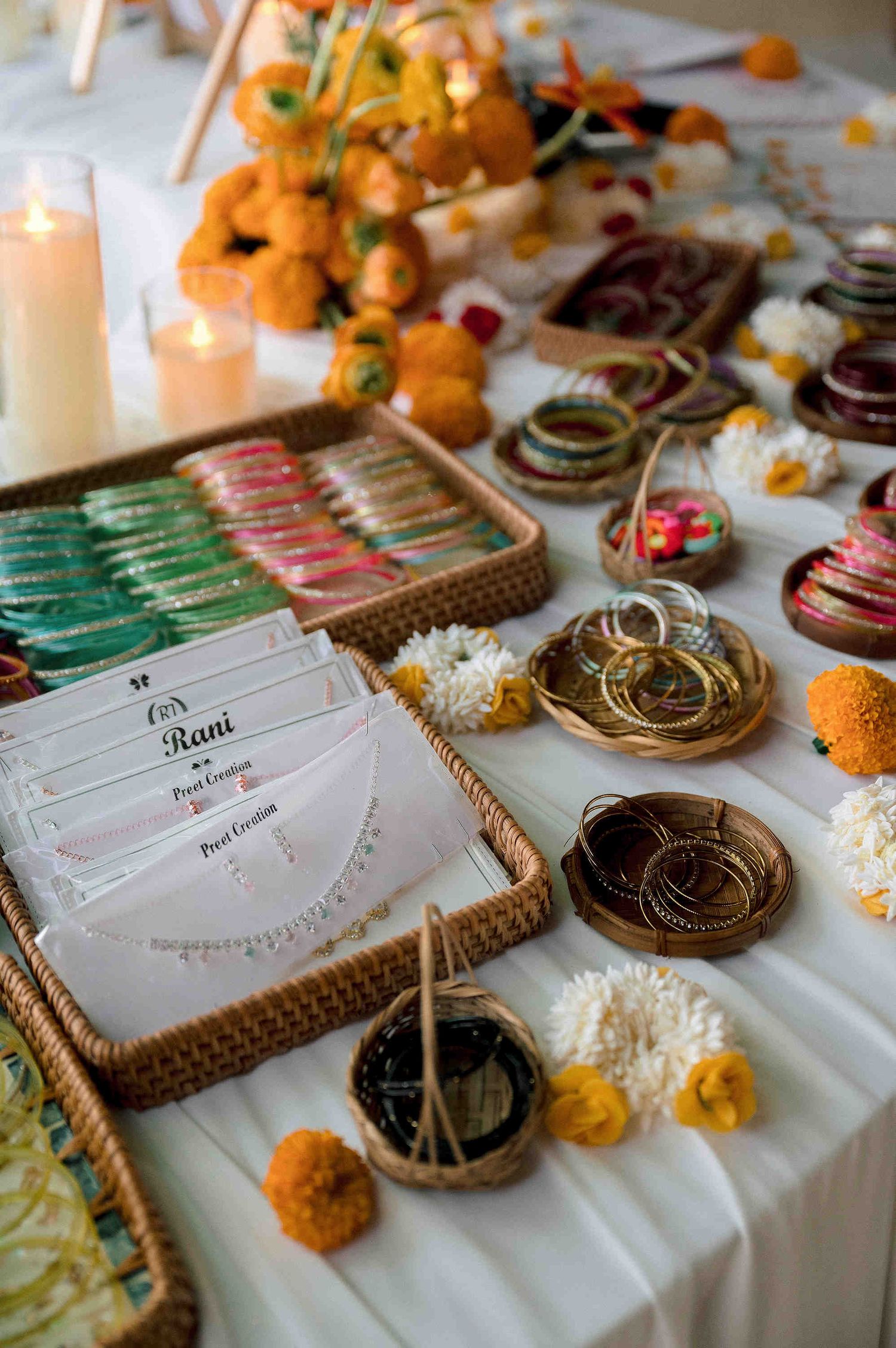 Indian wedding bangle station with marigold flowers, gold bangles, green chura, and personalized favor cards on white table.