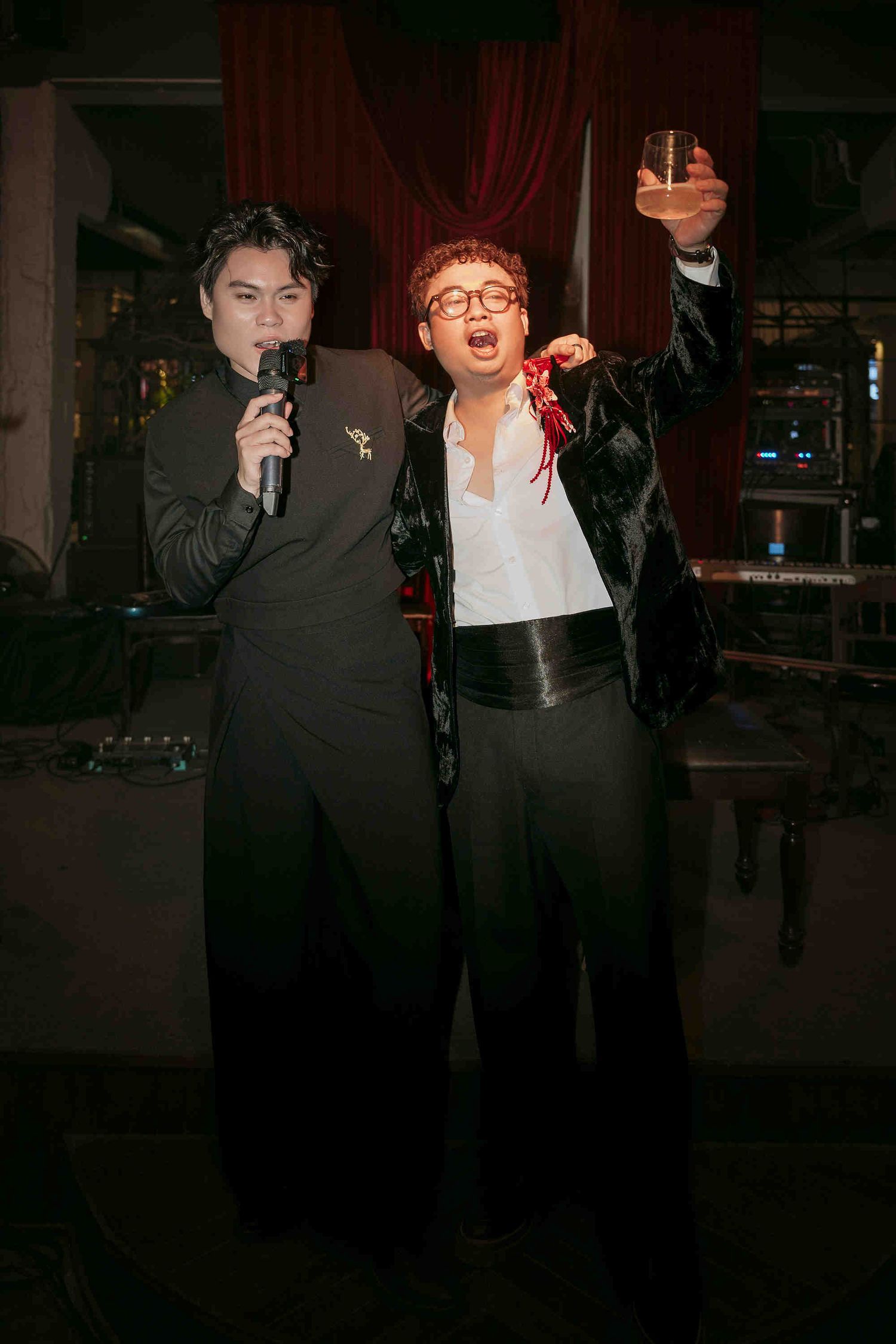 Two men perform at a party, one singing into a mic while the other raises a glass in celebration.