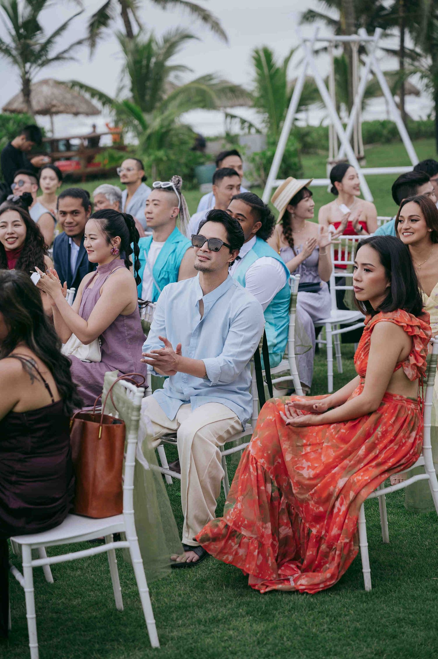Guests seated outdoors at a beachside wedding ceremony, dressed in colorful tropical attire on white chairs.