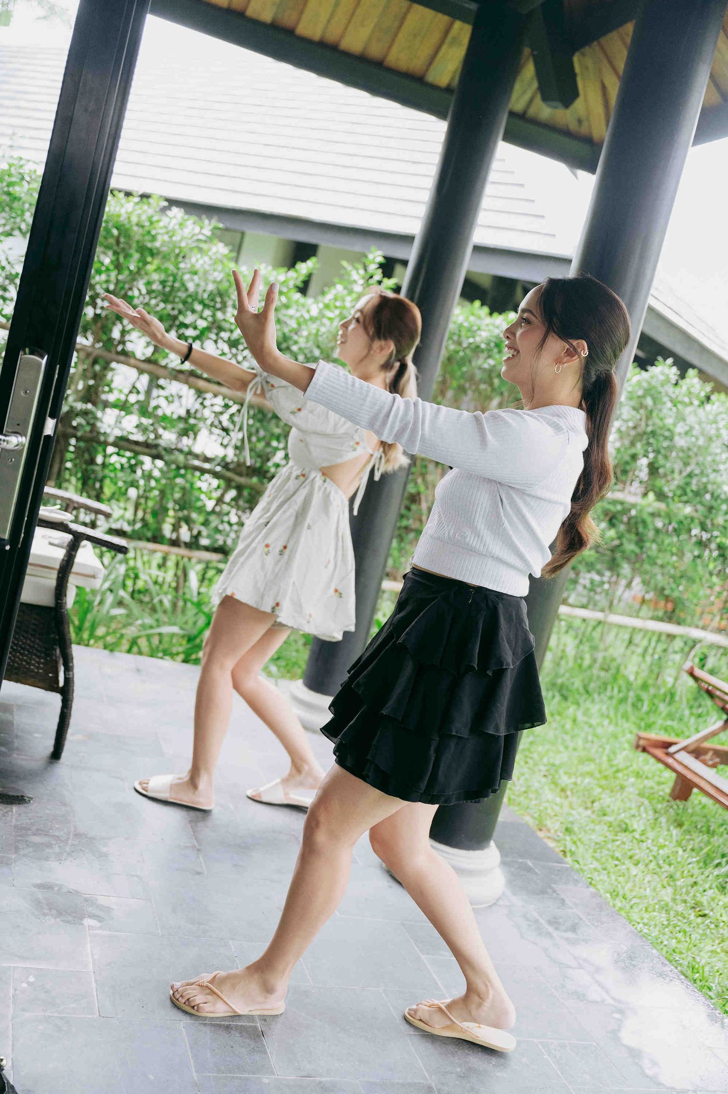 Two young women dancing playfully on a covered patio surrounded by lush greenery, one in white dress, one in black skirt.
