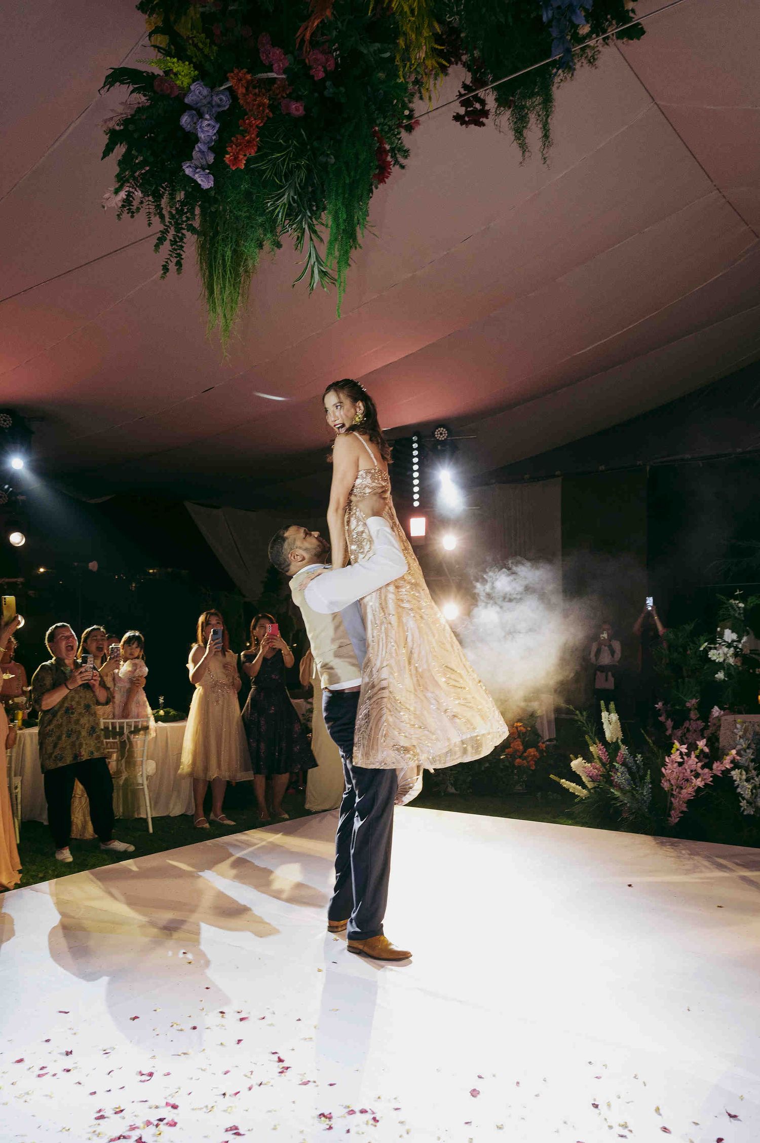 Groom lifting bride during first dance at wedding reception, surrounded by guests, floral ceiling installation above.