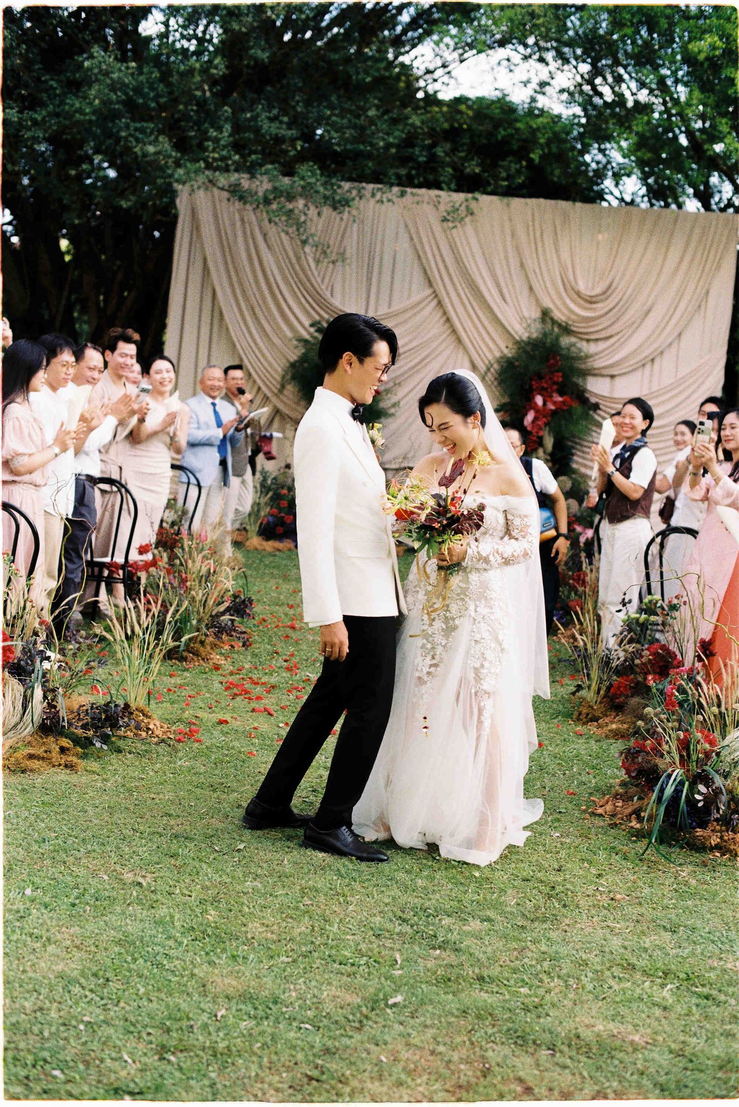 Newlywed couple walking down the aisle at an outdoor garden wedding ceremony with guests applauding.