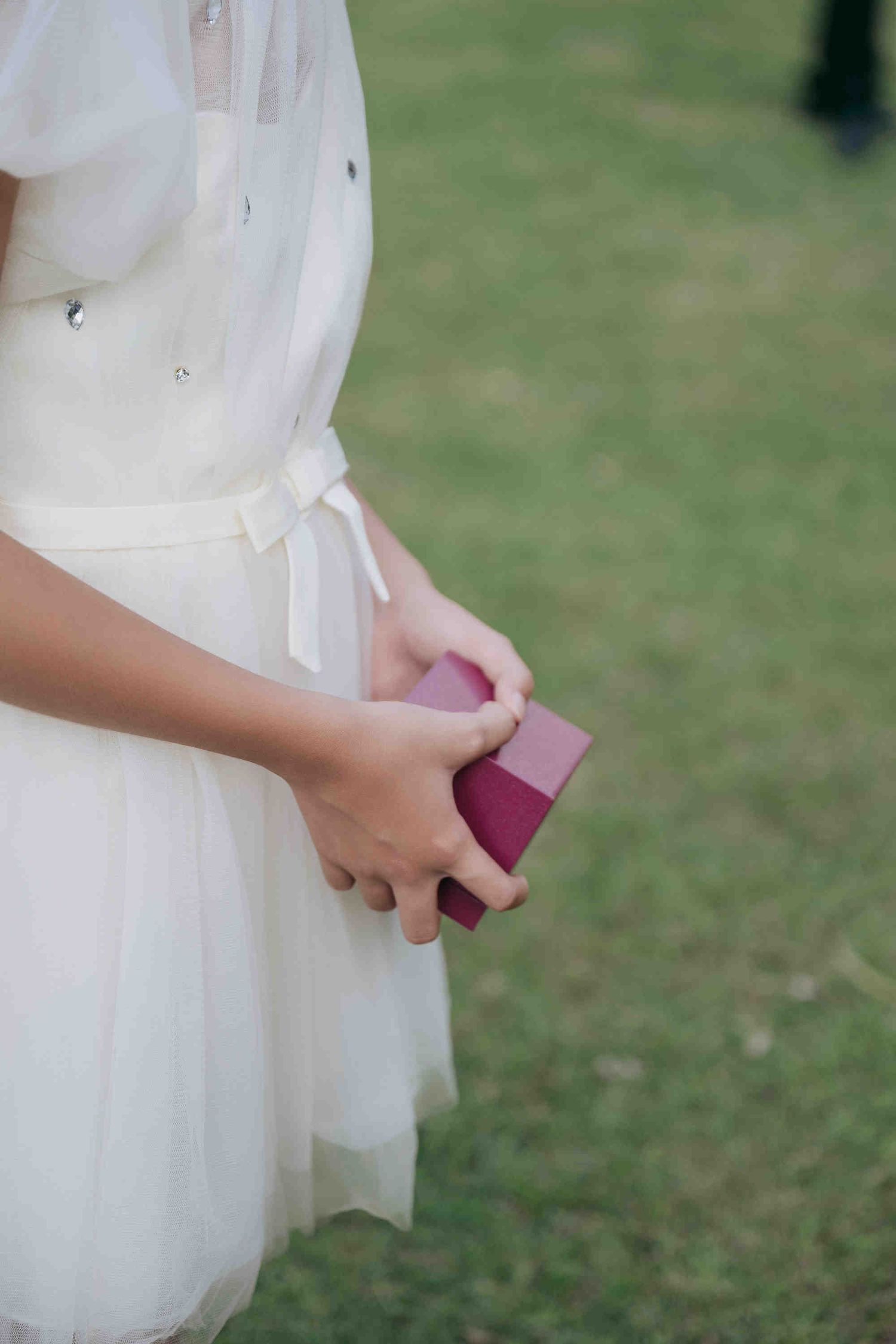Young girl in white dress holding a small burgundy gift box outdoors on green grass.