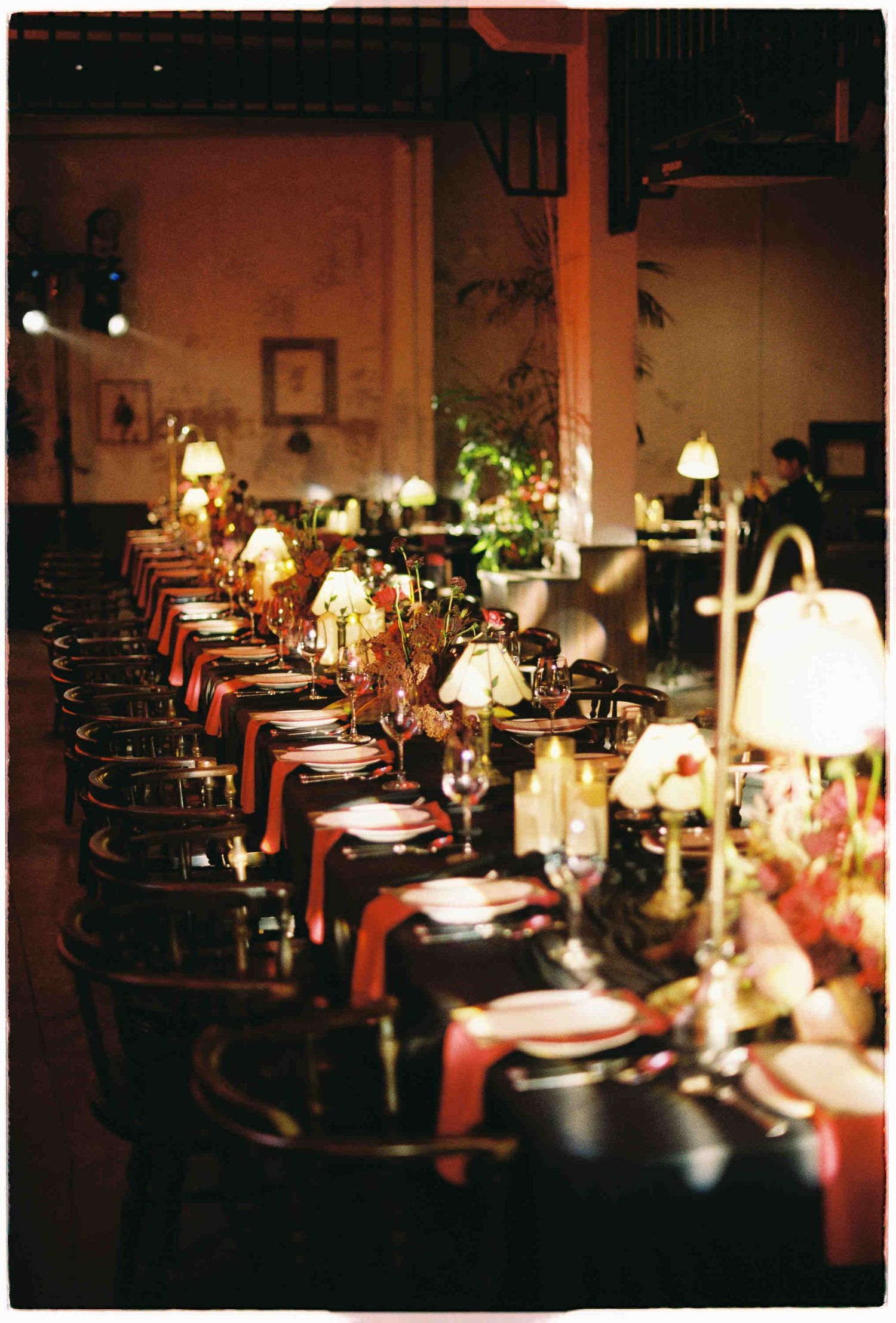 Elegant candlelit banquet table with dark linens, floral centerpieces, and small lamps in a warmly lit venue.