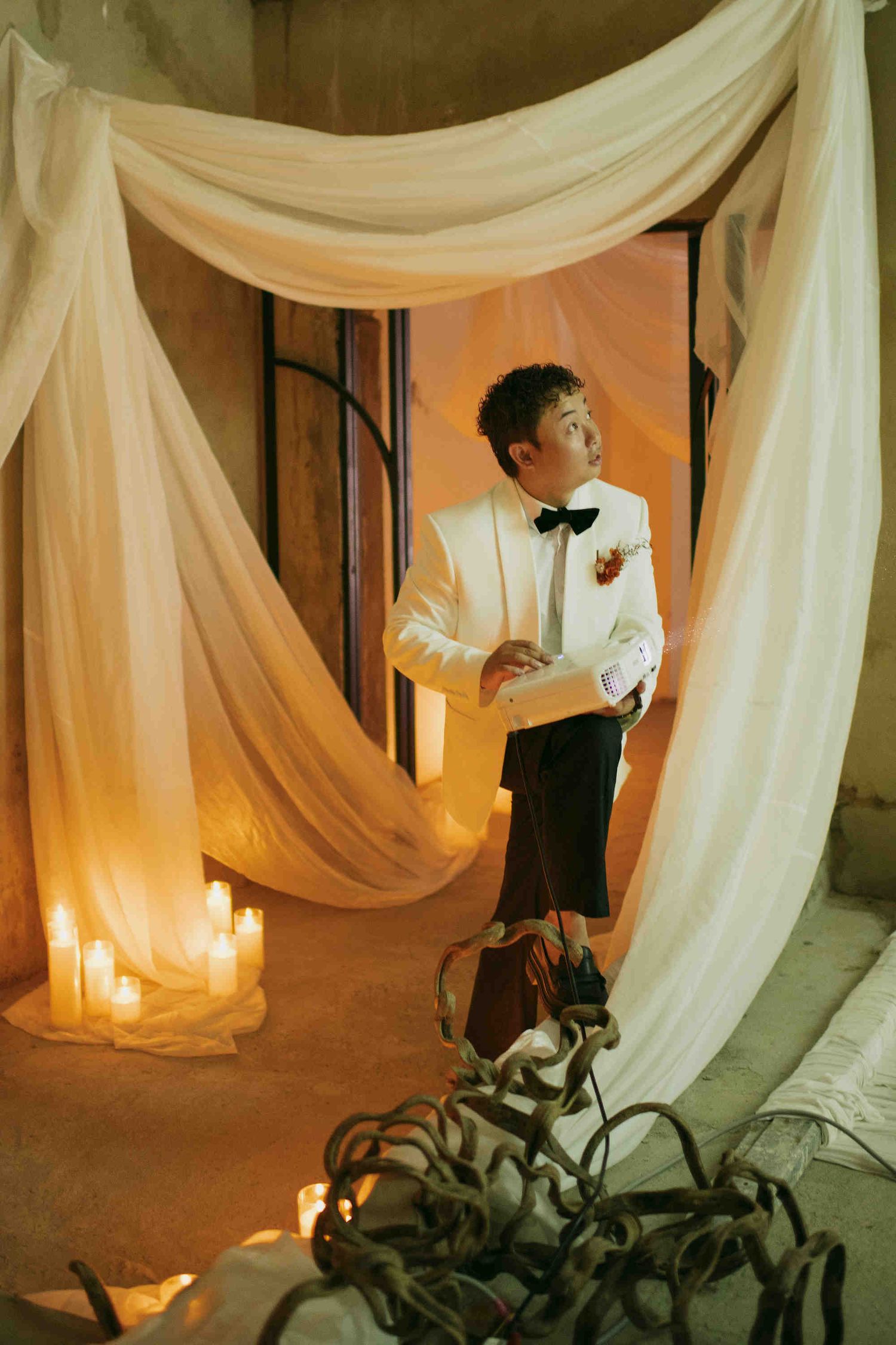 Elegant groom in white tuxedo jacket and bow tie standing in candlelit doorway with flowing white drapes at wedding venue.